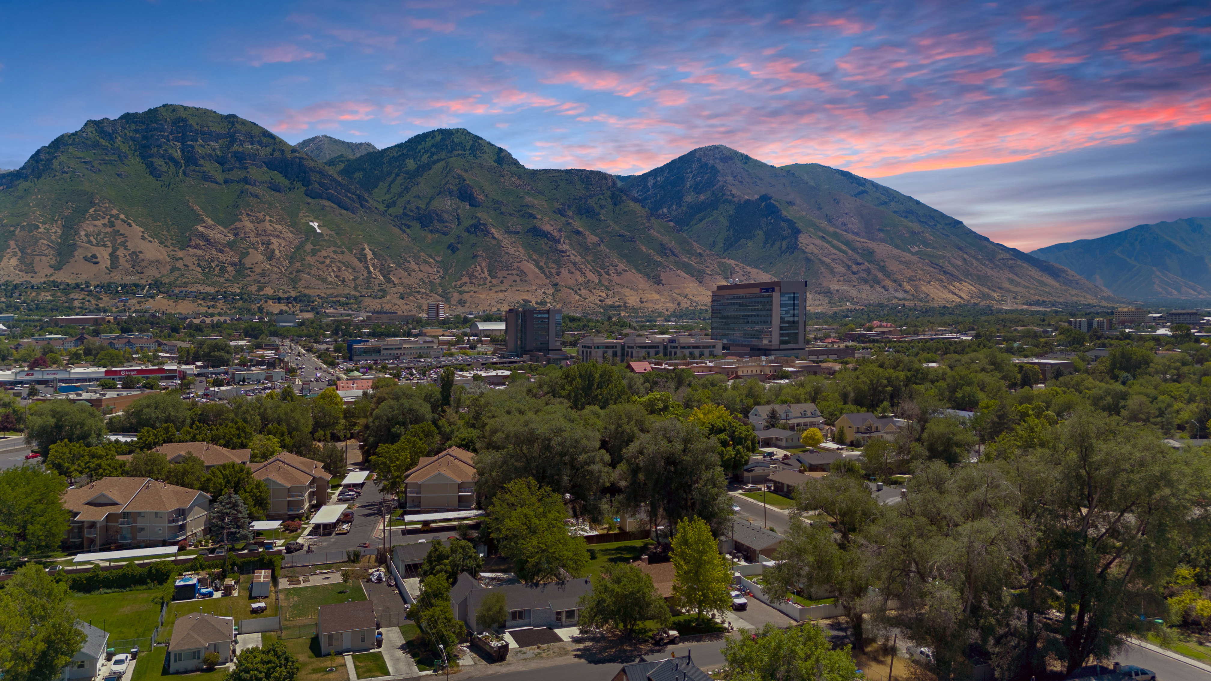 Expansive aerial perspective showcasing a mountainous backdrop, a verdant urban landscape with mature trees, a mix of residential structures, and commercial buildings