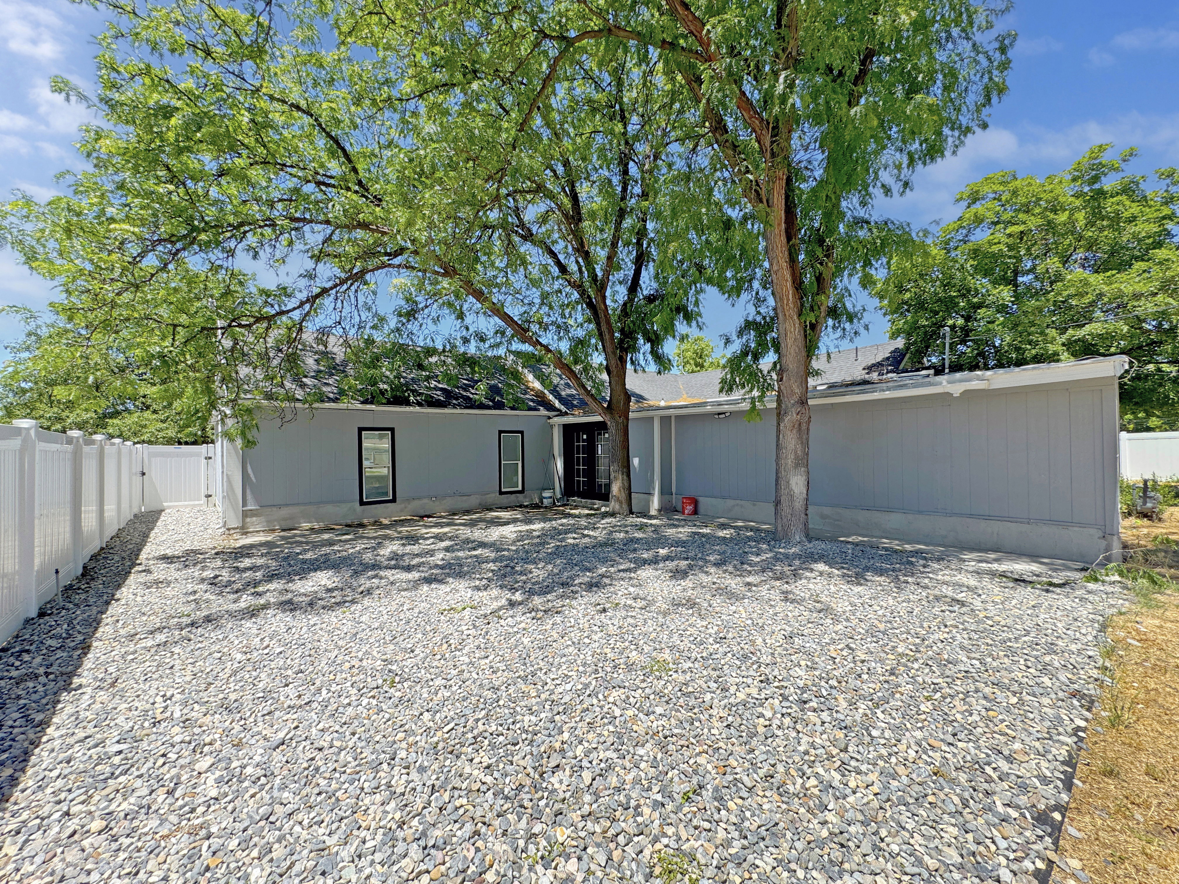 Rear exterior featuring light gray siding, a dark roof, and multiple mature trees providing natural shading