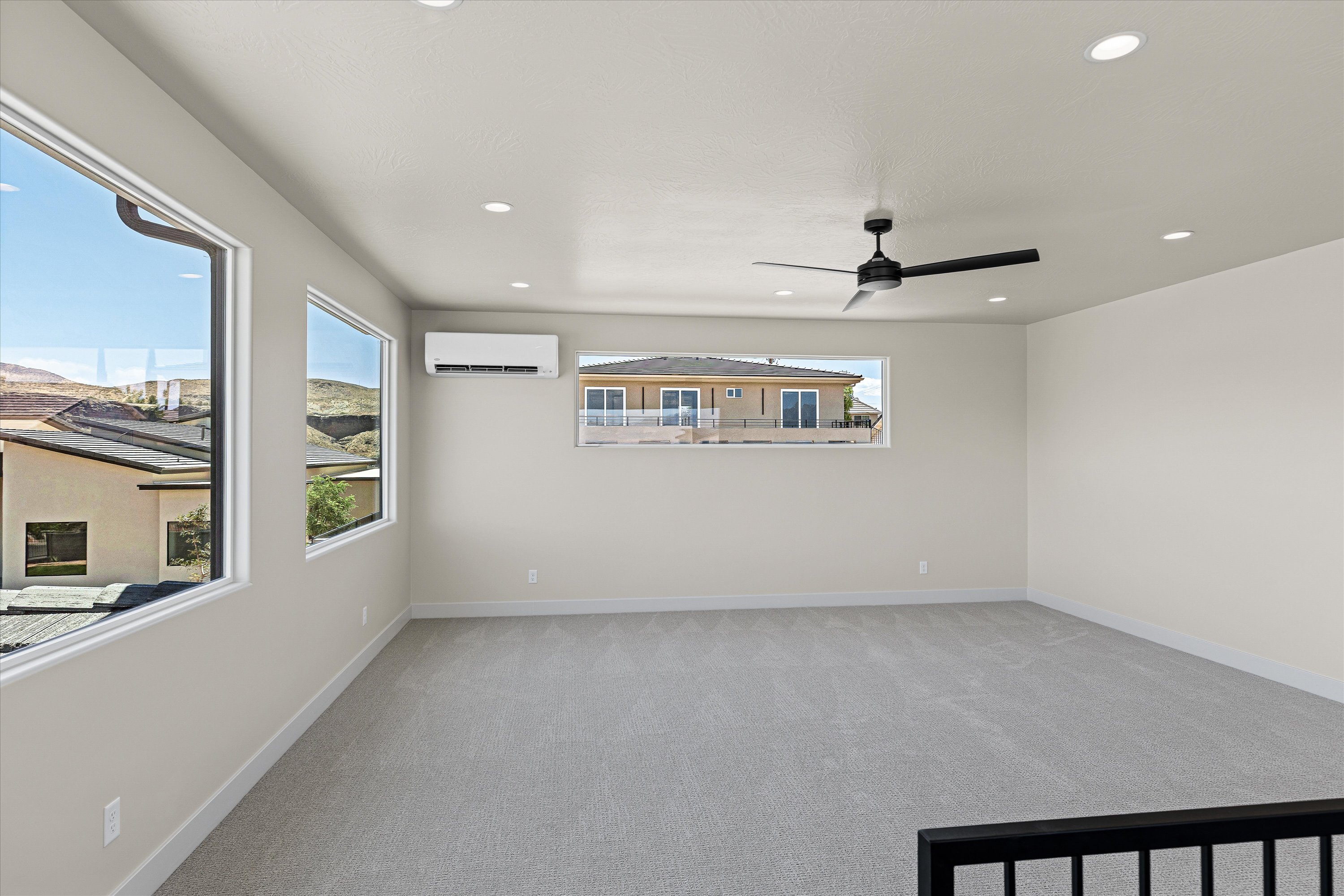 Spacious room featuring light gray carpeting, recessed ceiling lighting, a contemporary ceiling fan, multiple windows, and a wall-mounted climate control unit