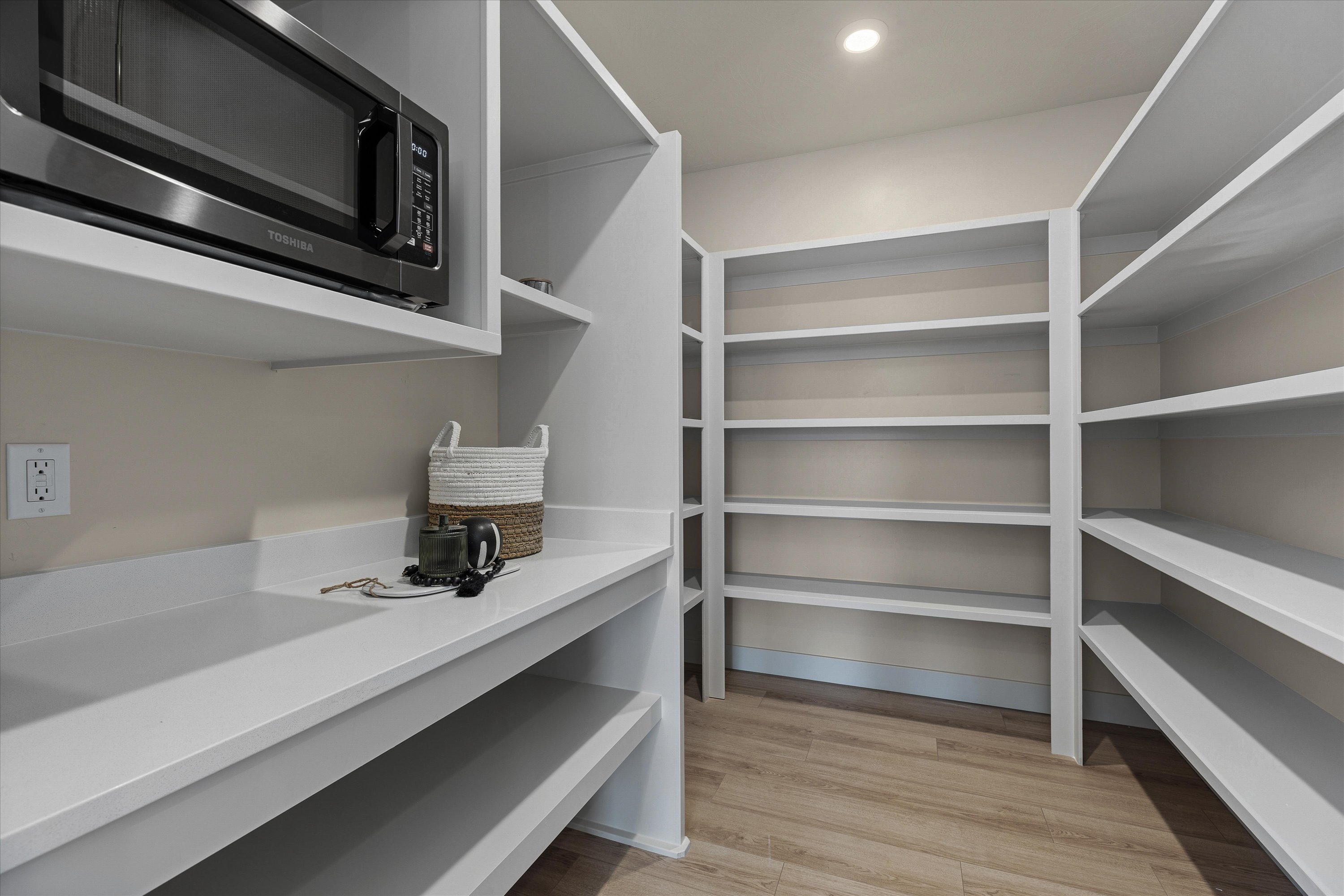 Thoughtfully designed pantry featuring extensive shelving, a built-in microwave, white countertops, wood-finish flooring, and recessed lighting
