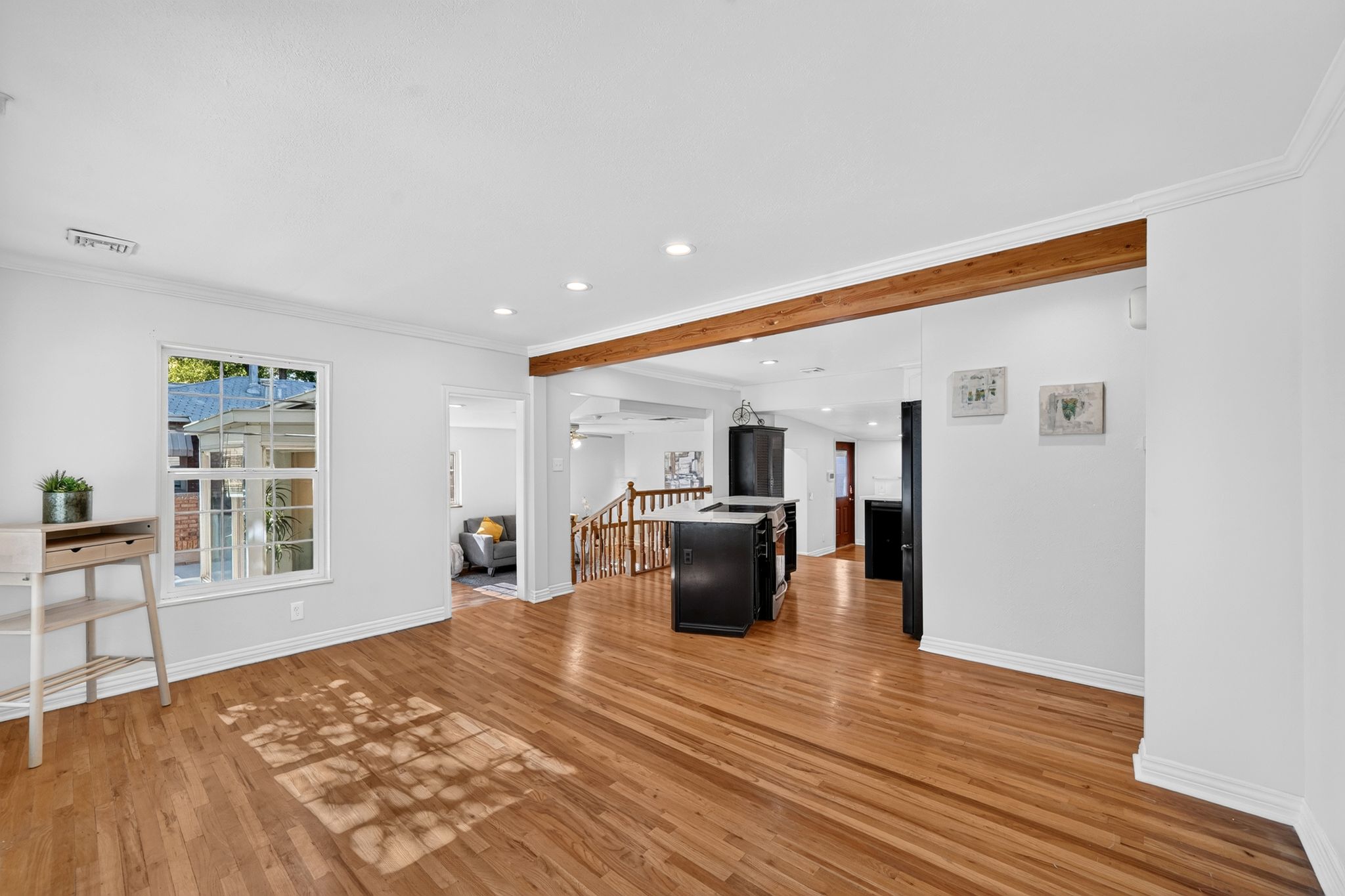 Open-concept living space with warm wood-finish flooring, white walls, recessed lighting, and a prominent wood beam accent