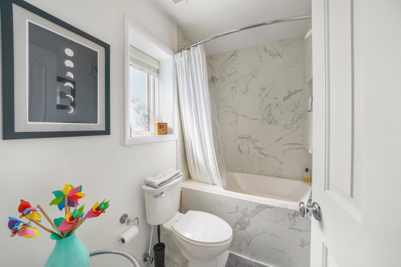 Bathroom featuring a white bathtub with marble-look tile surround, a white toilet, and a window with a white blind