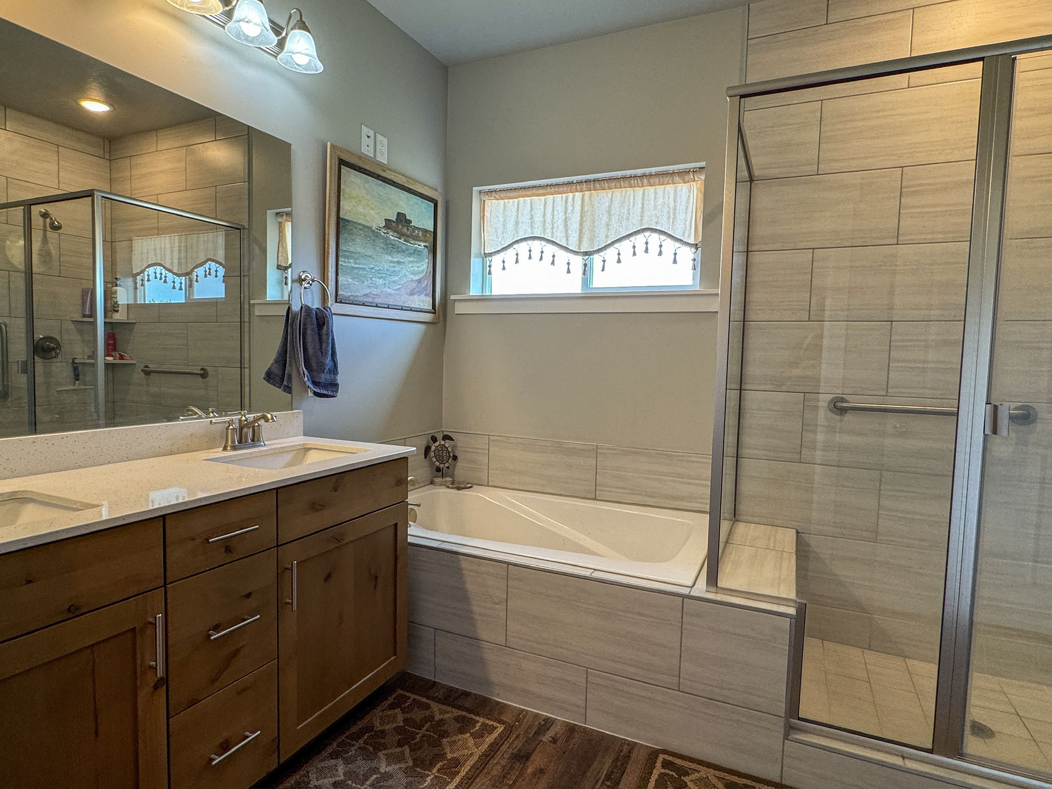 Bathroom featuring a dual vanity with light-toned countertops, a glass-enclosed shower with tile surround, a built-in bathtub with matching tile surround, and wood-finish flooring