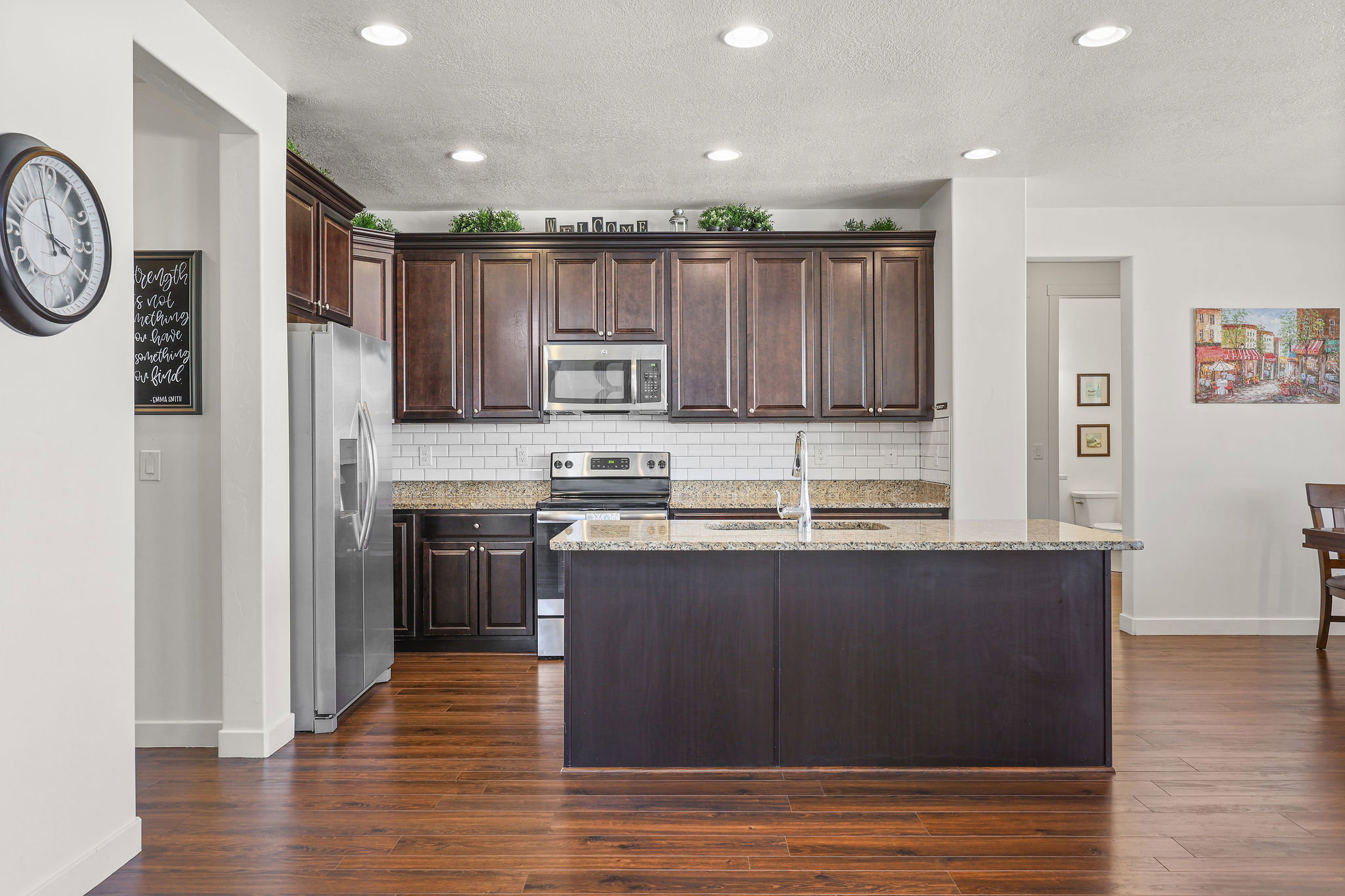 Kitchen featuring dark wood cabinetry, a central island with a sink, stainless steel appliances, granite-finish countertops, and a white subway tile backsplash