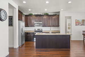 Kitchen featuring dark wood cabinetry, a central island with a sink, stainless steel appliances, granite-finish countertops, and a white subway tile backsplash