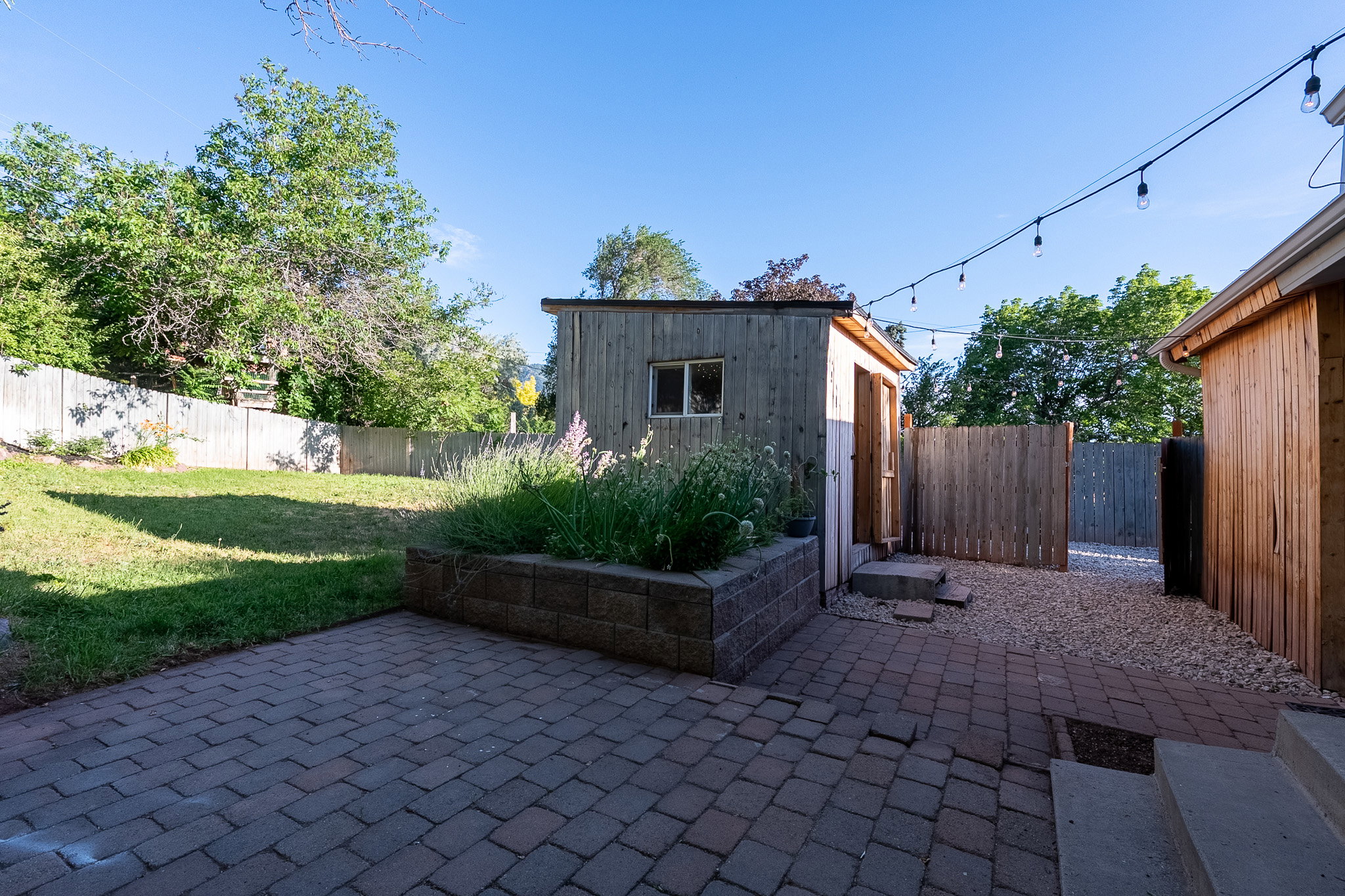 Paved patio area with a raised garden bed, gravel pathway, and a wooden storage shed