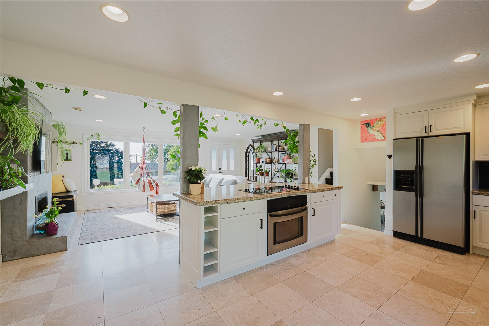 Open-concept interior featuring light-toned tile flooring, recessed lighting, a kitchen island with built-in oven and cooktop, white cabinetry, and stainless steel refrigerator
