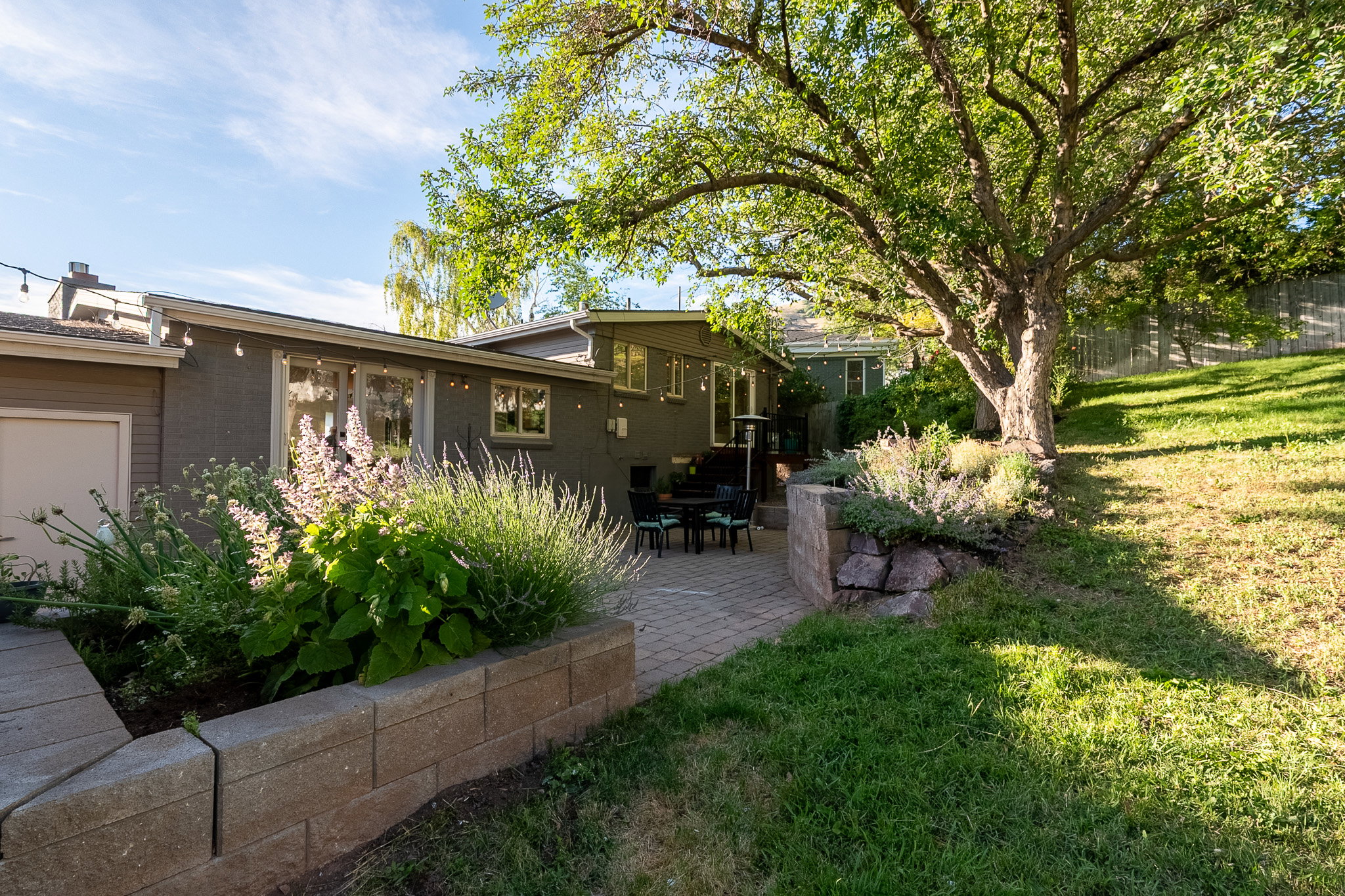 Expansive backyard featuring a paver patio with a stone retaining wall, mature trees, and established plantings