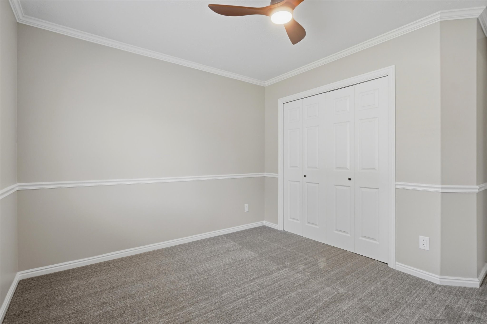 Neutral-toned room featuring a ceiling fan with integrated lighting, bi-fold closet doors, crown molding, chair rail molding, and grey carpet flooring