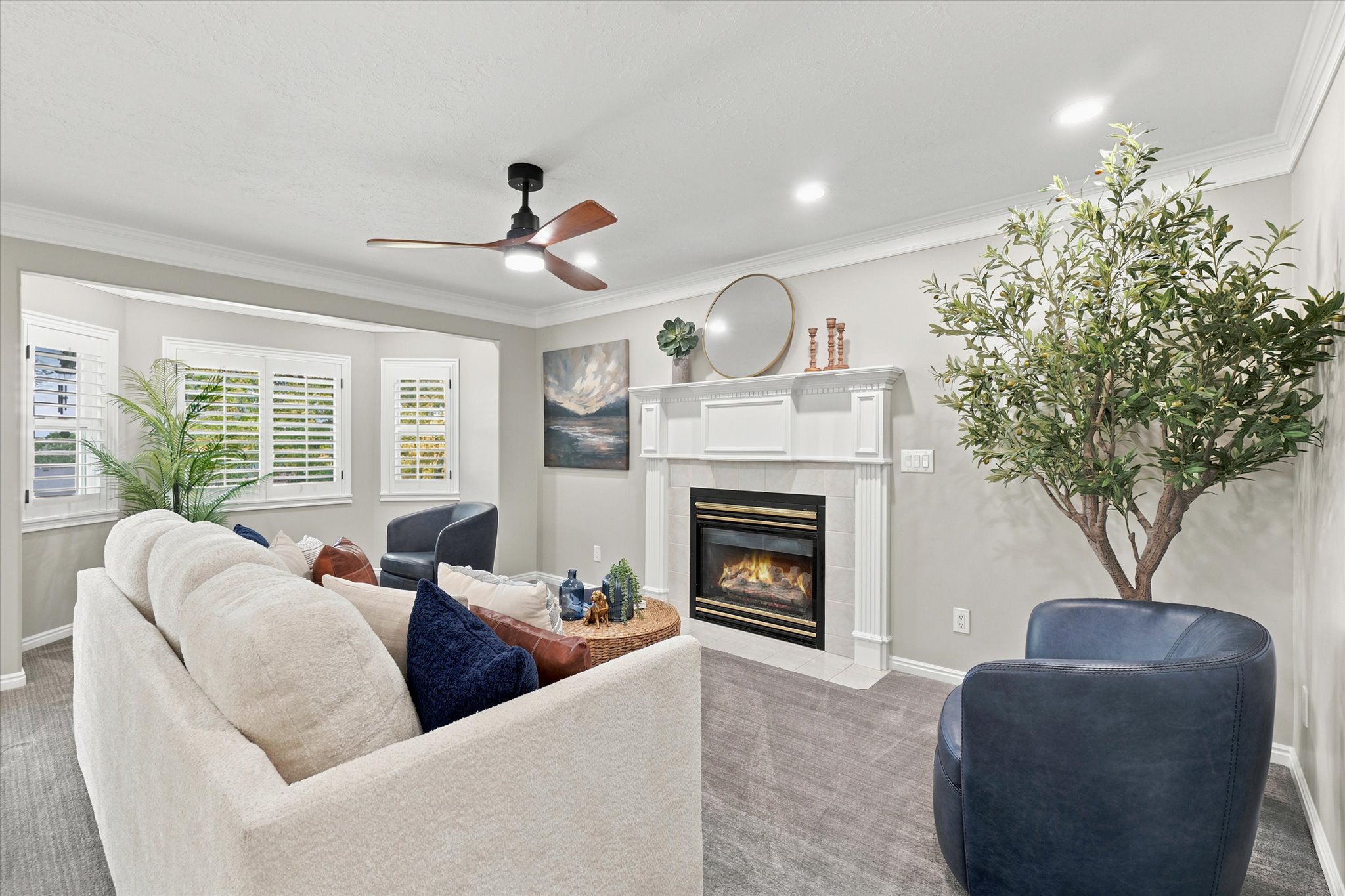 Living area featuring a gas fireplace with a white mantel, bay windows with plantation shutters, a ceiling fan with integrated lighting, recessed lighting, and crown molding