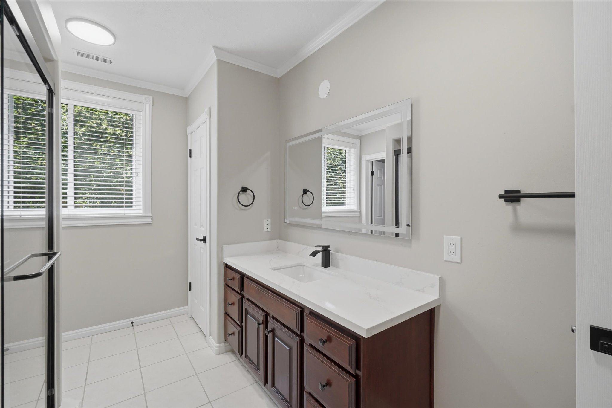Bathroom featuring a rich wood-finish vanity with a white countertop and integrated sink, a frameless glass shower enclosure, a rectangular wall-mounted mirror, and tile flooring