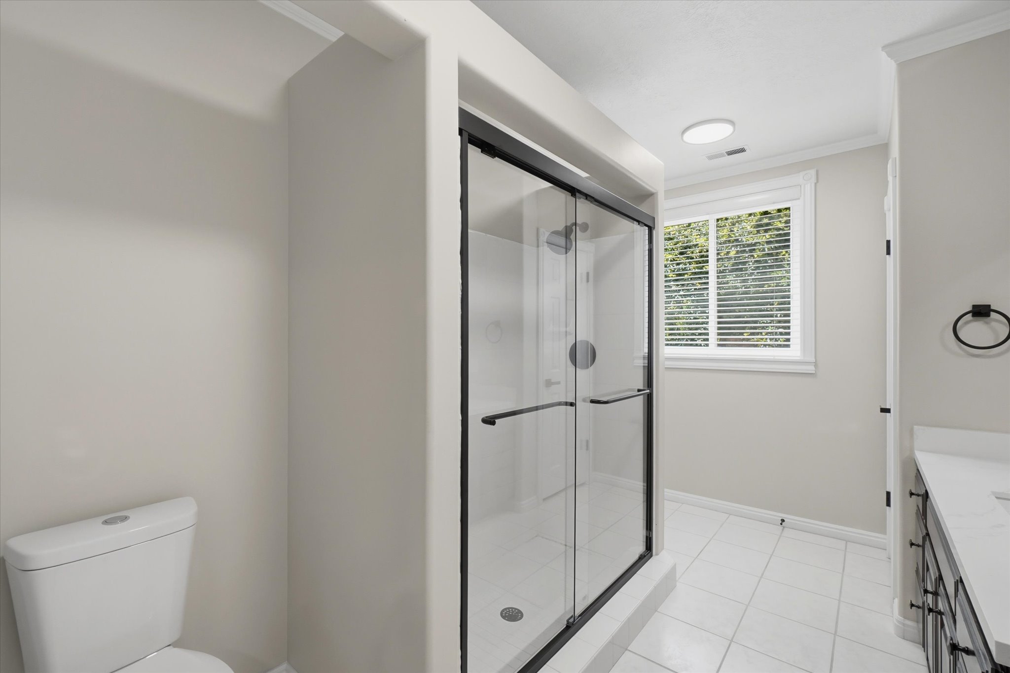 Full bathroom featuring a frameless glass shower enclosure with matte black trim, white square floor tiles, and a window with blinds
