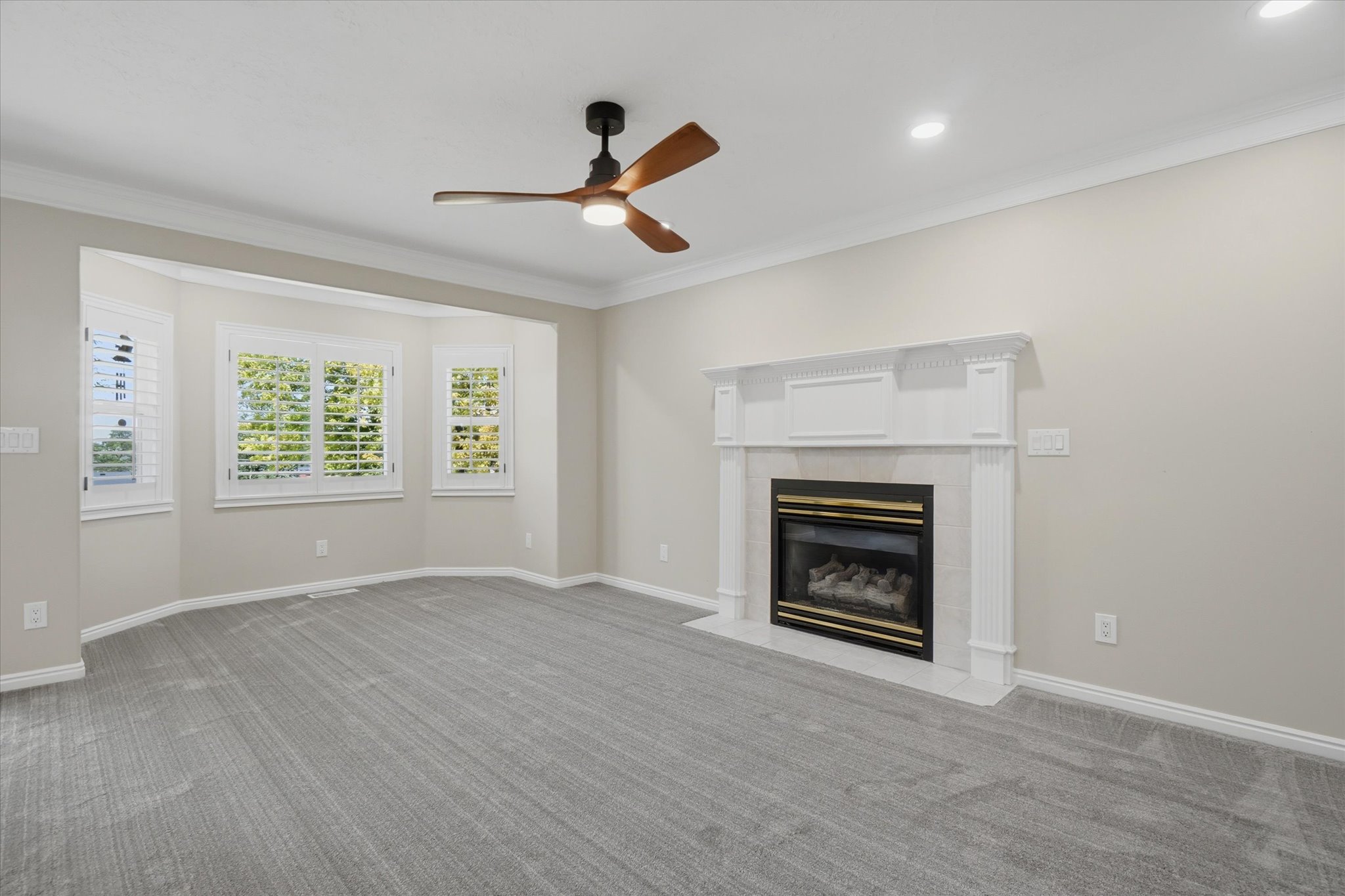 Living area featuring a gas fireplace with a decorative mantel, bay windows with plantation shutters, a ceiling fan with wood-finish blades, recessed lighting, and gray carpet flooring