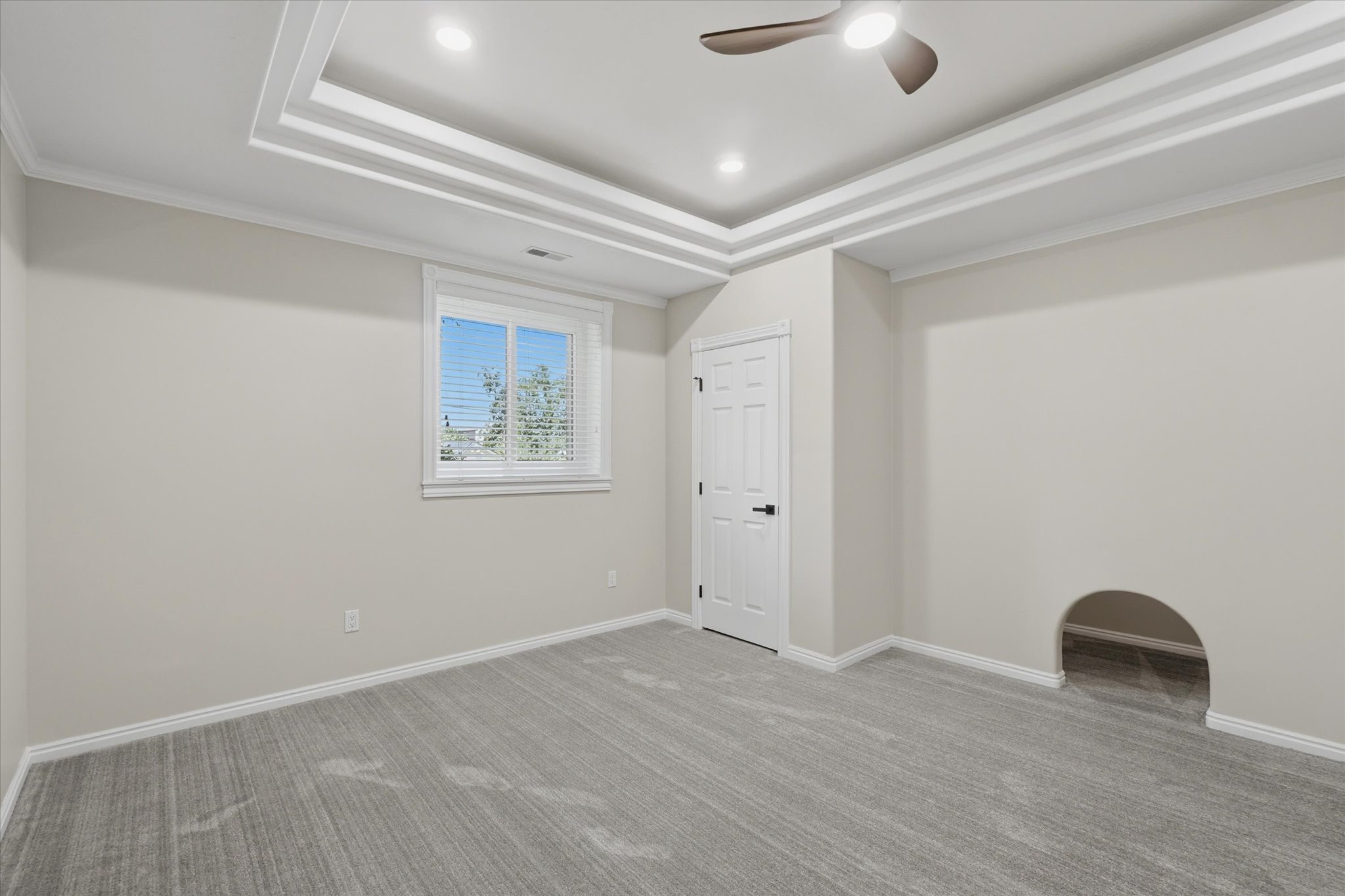 Recessed tray ceiling with integrated lighting, modern ceiling fan, gray carpet flooring, white trim, and a window with blinds