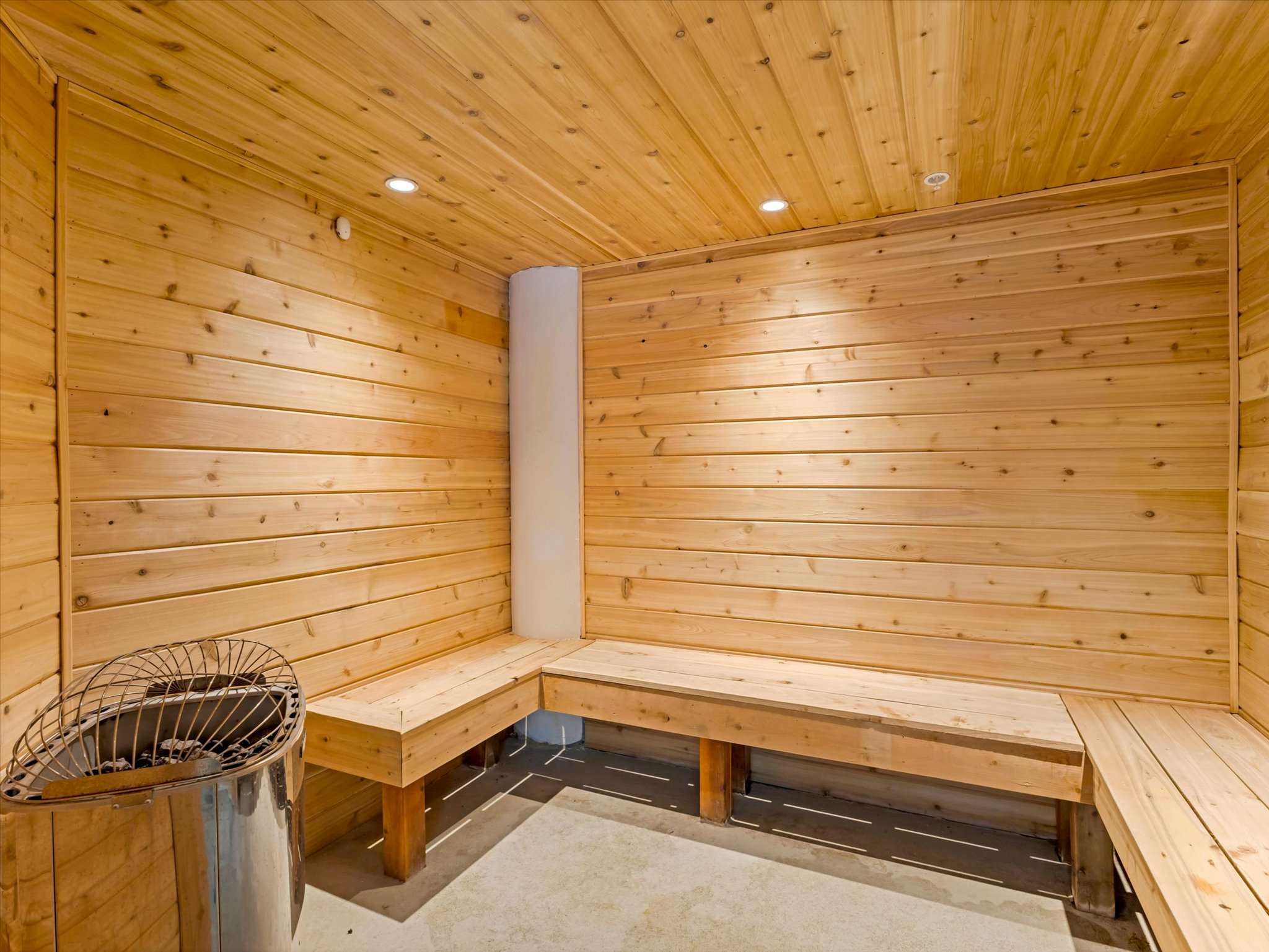Wood-paneled sauna featuring built-in benches, recessed lighting, concrete flooring, and a stainless steel heater