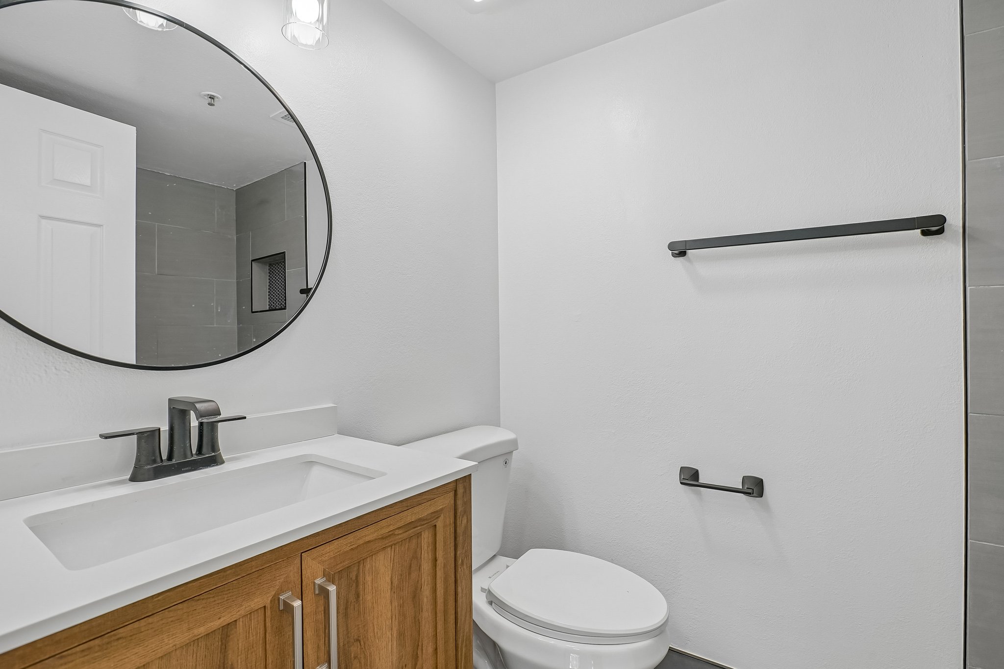 Bathroom featuring a wood-finish vanity with a white countertop and integrated sink, matte black faucet, and a round black-framed mirror