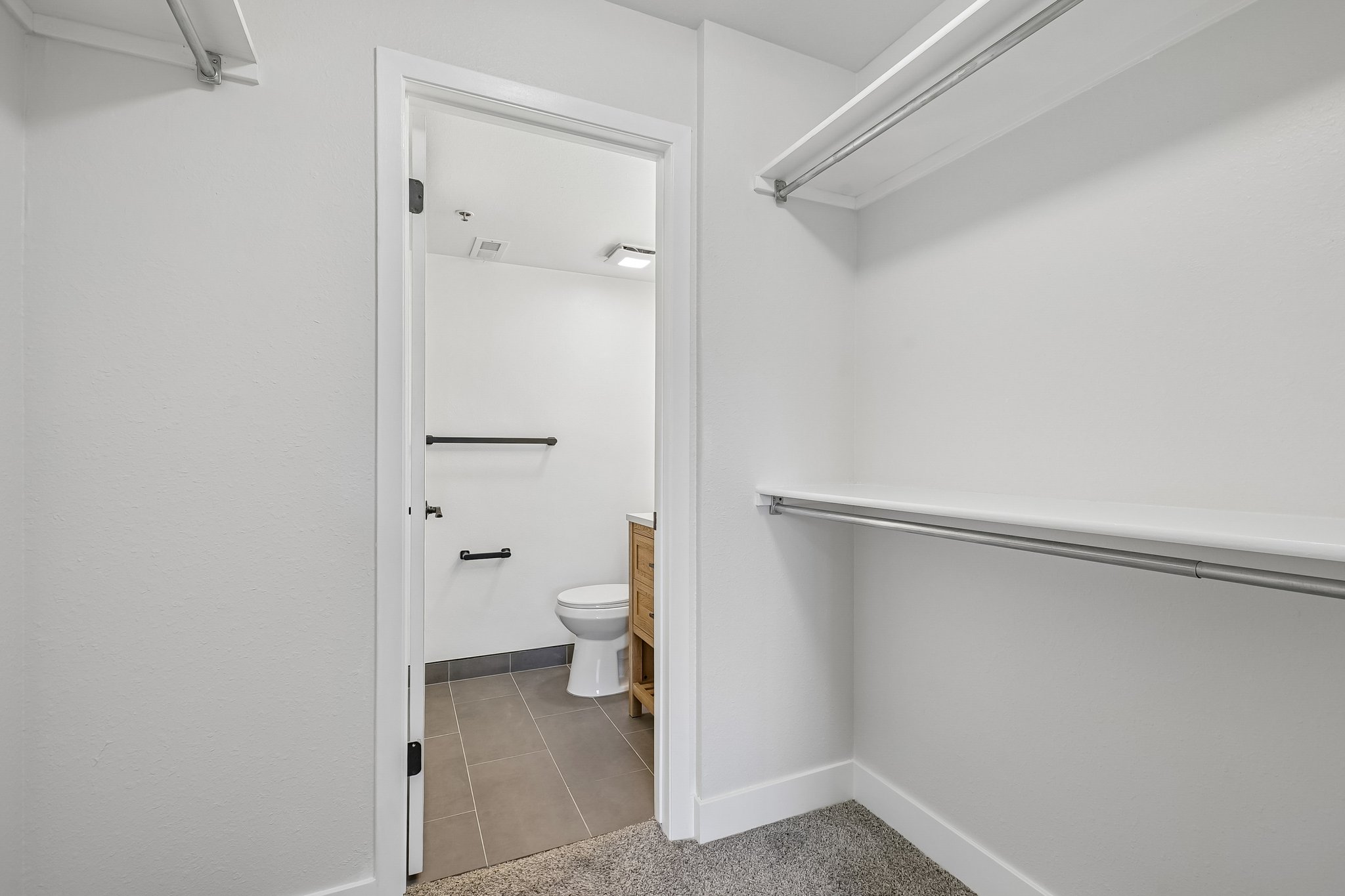 Walk-in closet with carpet flooring, a top shelf, and a lower hanging rod, opening to a bathroom with tile flooring, a toilet, and a wood-finish vanity with a vessel sink