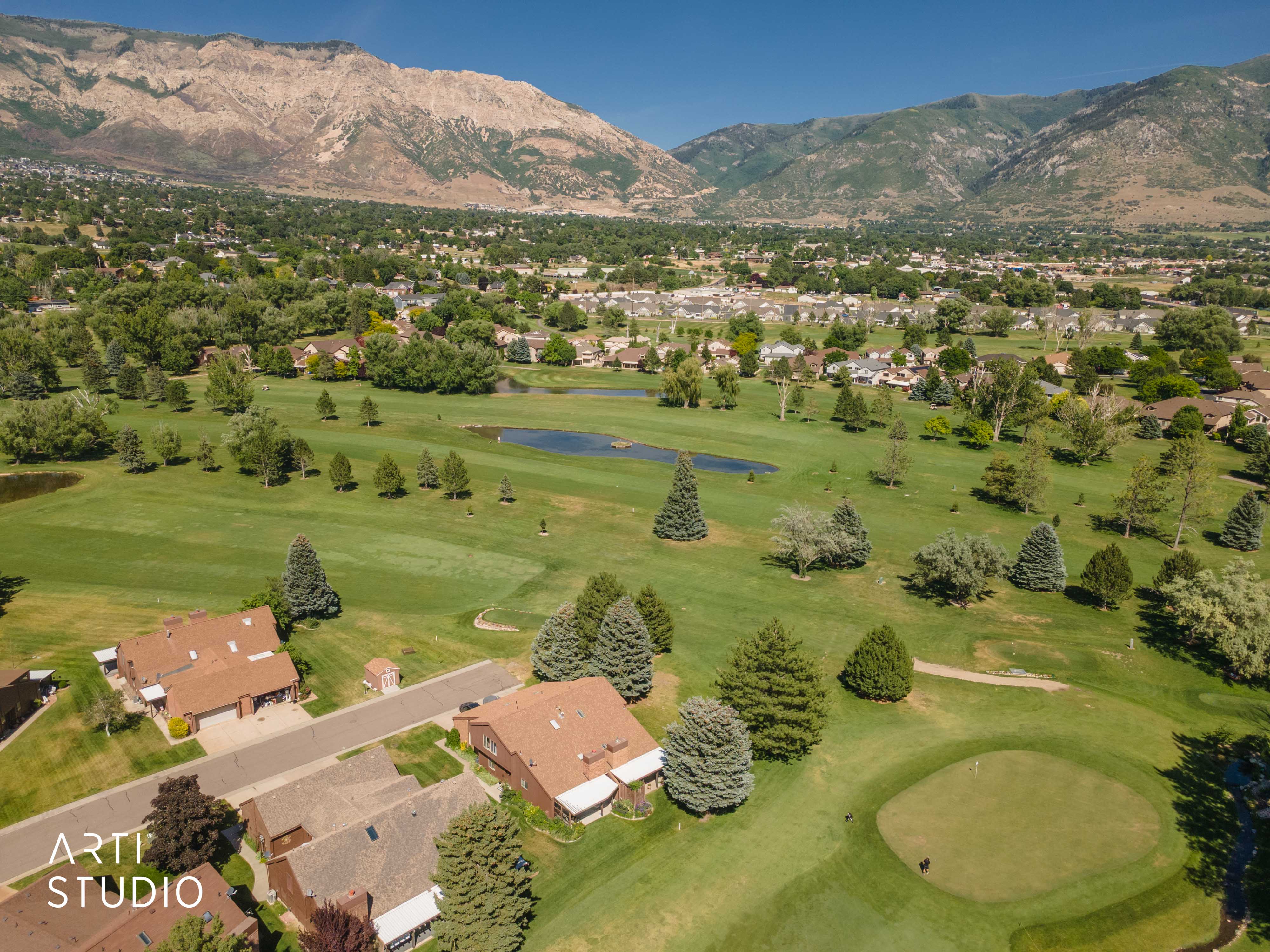 Expansive golf course setting featuring manicured greens, mature trees, and water features, all against a dramatic mountain backdrop