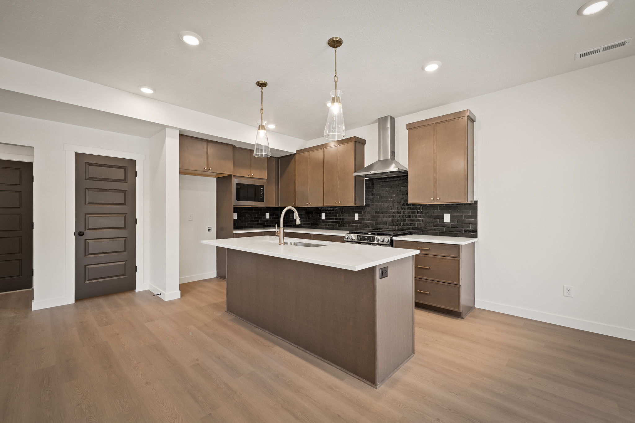 Contemporary kitchen featuring a large central island with a solid surface countertop and sink, dark wood-finish cabinetry, black subway tile backsplash, stainless steel range hood, and recessed lighting