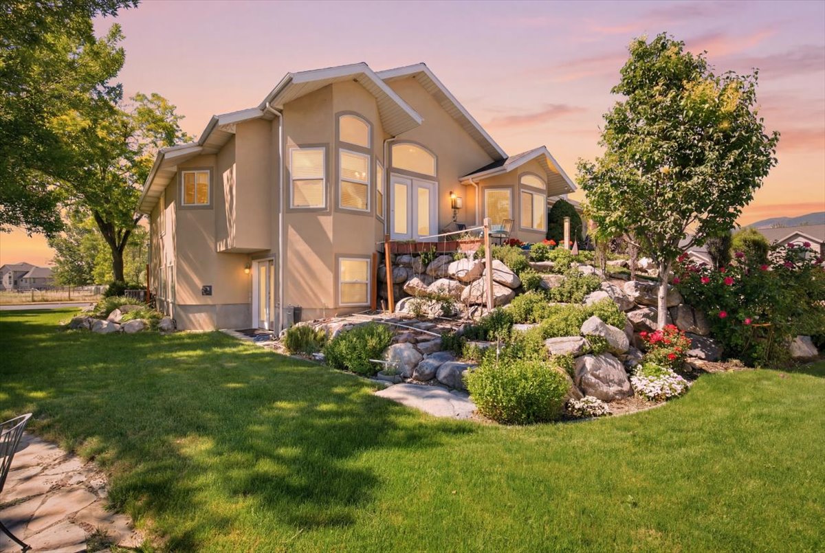 Exterior stucco residence featuring multiple windows, a stone retaining wall with plantings, a mature tree, and a grassy lawn