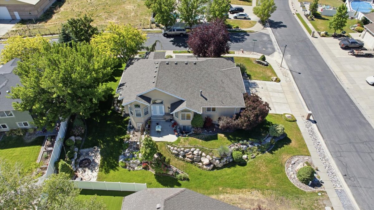 Contemporary residence featuring a light-toned exterior, a multi-car driveway, a stone-lined garden bed, a fire pit area, and a green lawn with mature trees