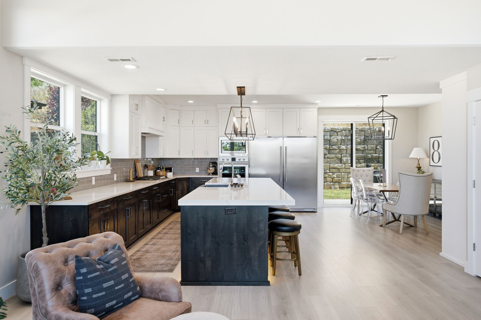 Open-concept kitchen featuring light wood-finish flooring, a central island with bar seating, stainless steel appliances, white upper cabinetry, and dark lower cabinetry with a tile backsplash