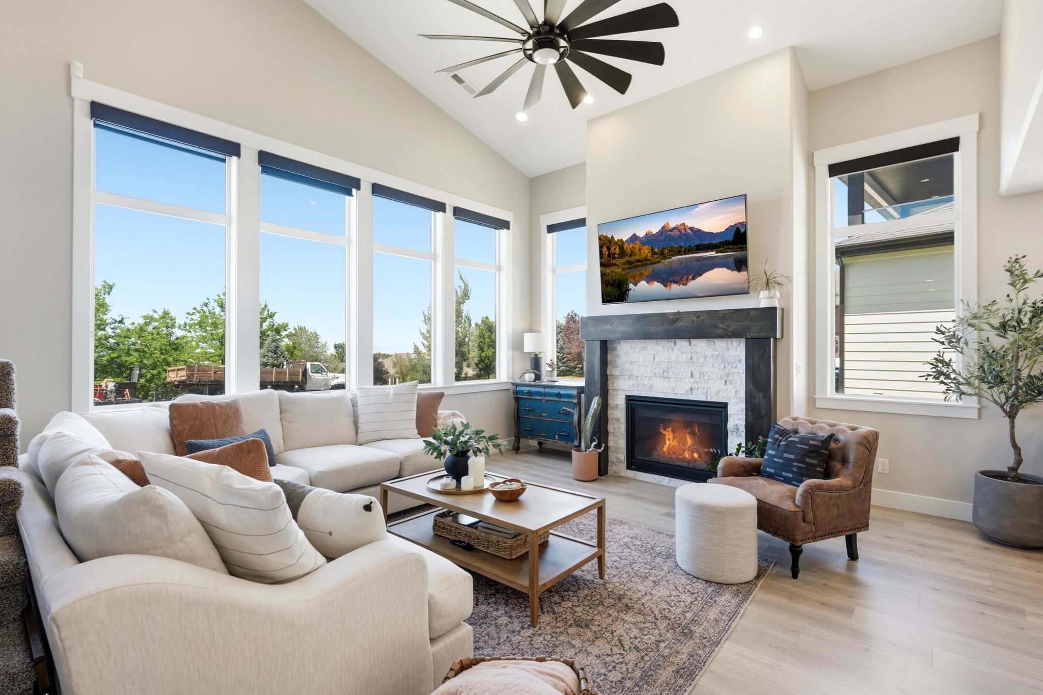 Vaulted ceiling living room featuring a gas fireplace with a white brick surround and dark wood mantel, large windows, wood-finish flooring, and a ceiling fan