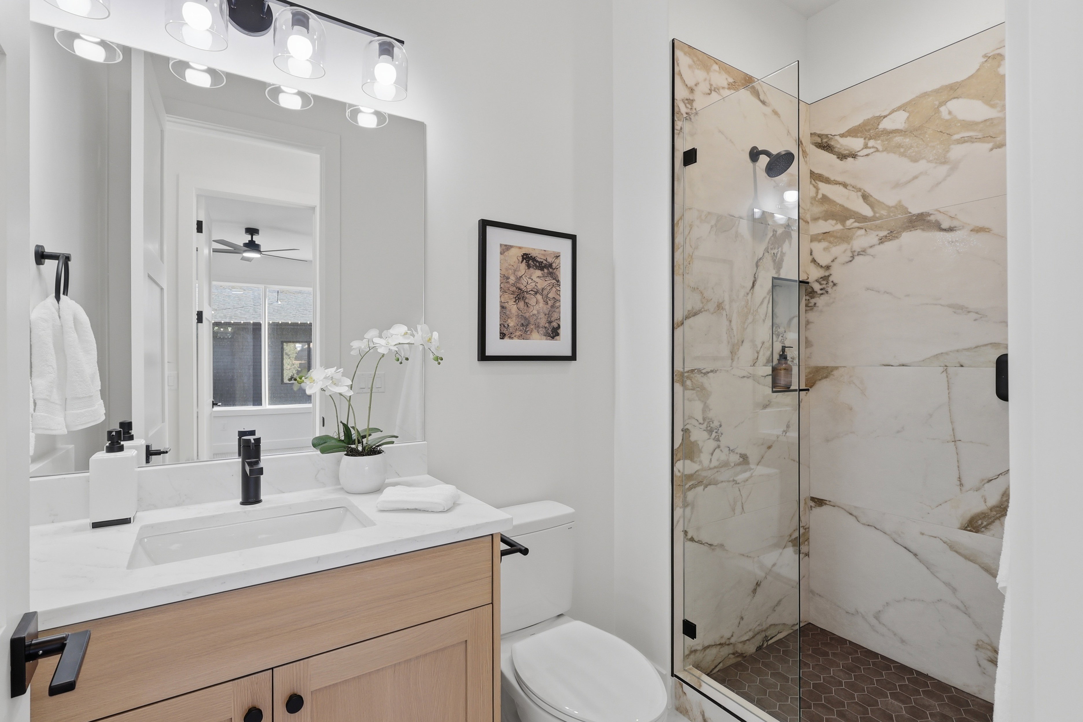 Bathroom featuring a frameless glass shower enclosure with floor-to-ceiling marbled tile surround and hexagon tile flooring, a wood-finish vanity with a white countertop and integrated sink, and a contemporary black fixture package
