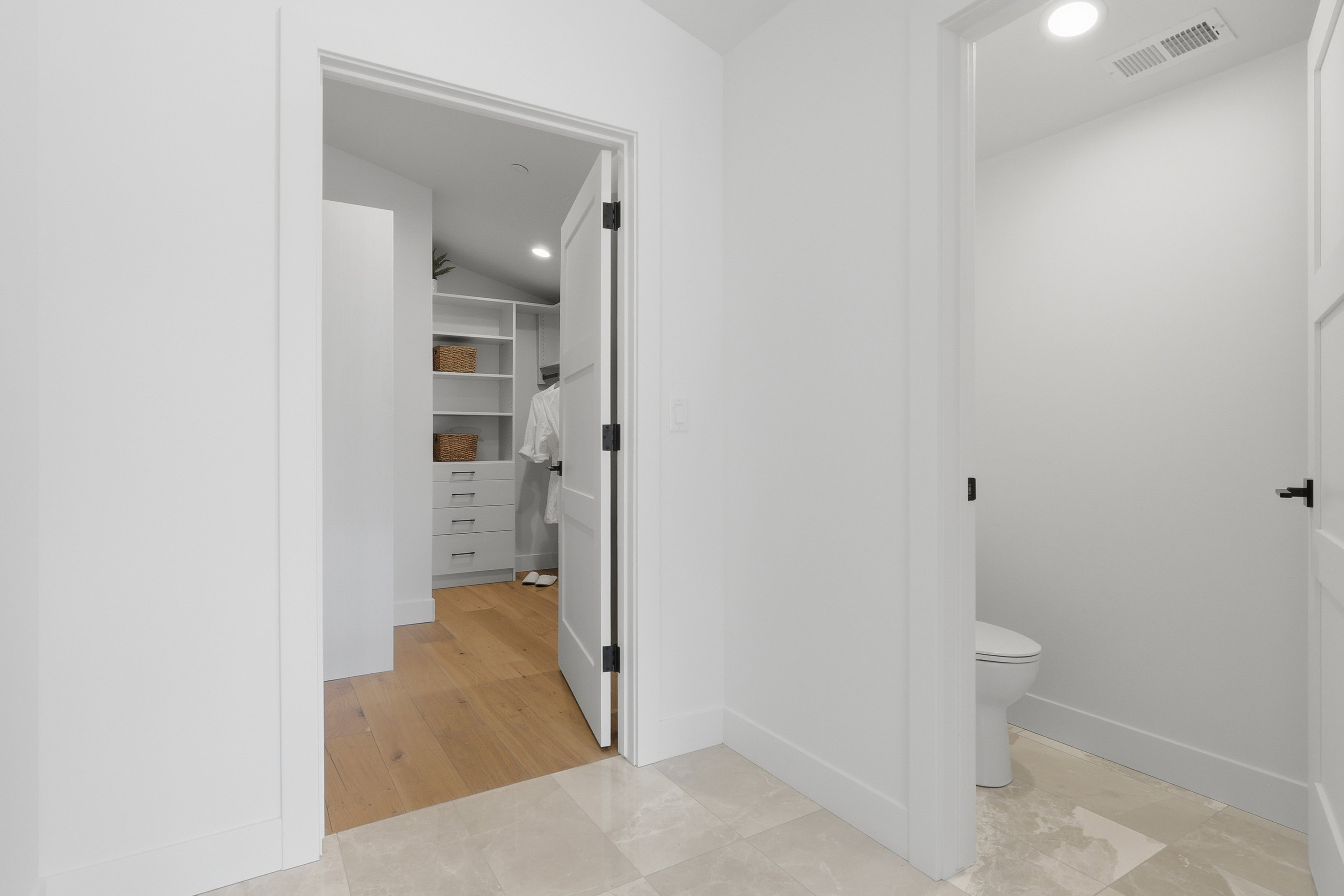 Hallway featuring light-toned tile flooring and white walls, leading to a closet with wood-finish flooring, built-in shelving, and drawers