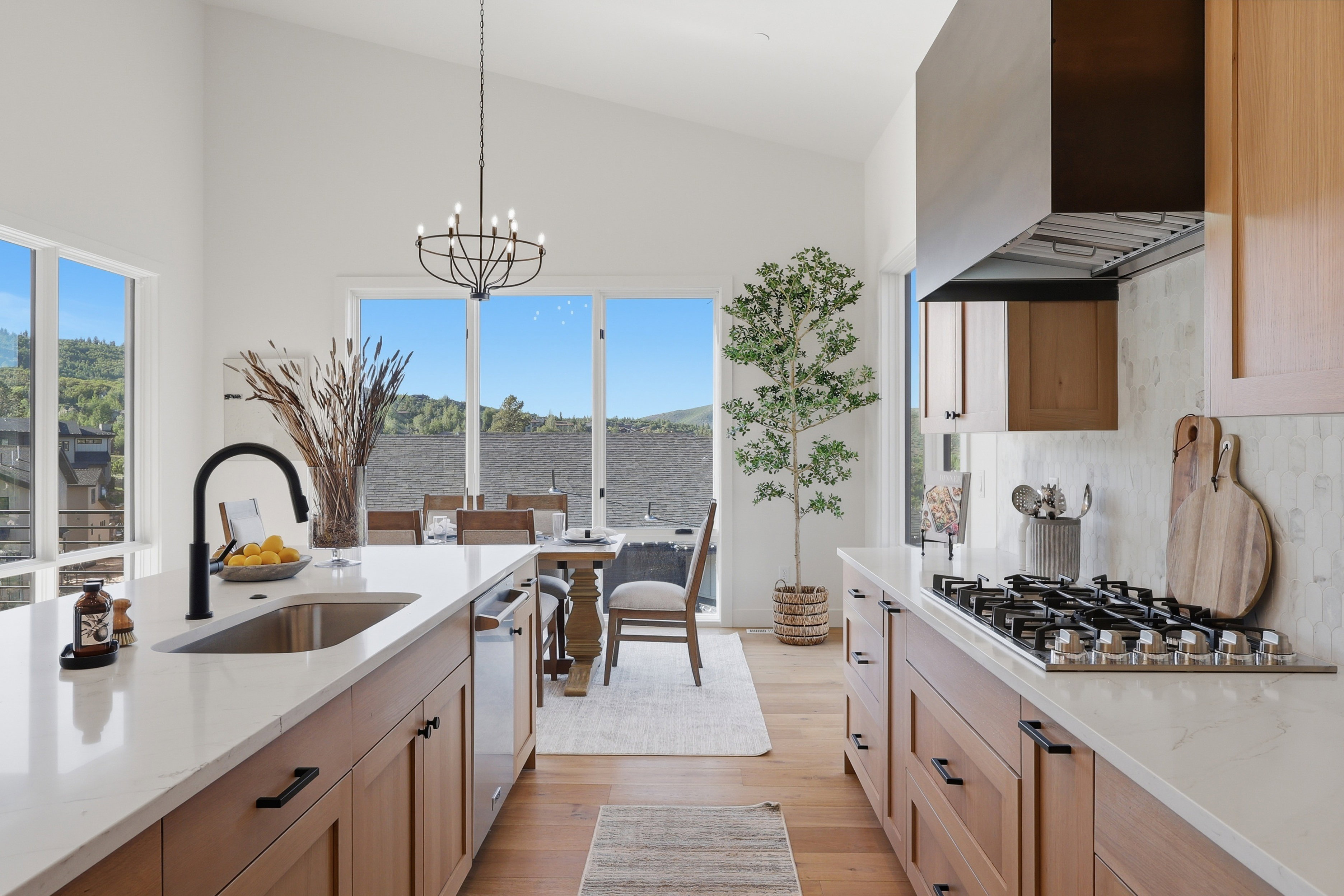 Open-concept kitchen and dining area featuring vaulted ceilings, light wood-finish cabinetry, a large kitchen island with an undermount sink and black faucet, a gas range with a stainless steel vent hood, and extensive natural light from multiple windows