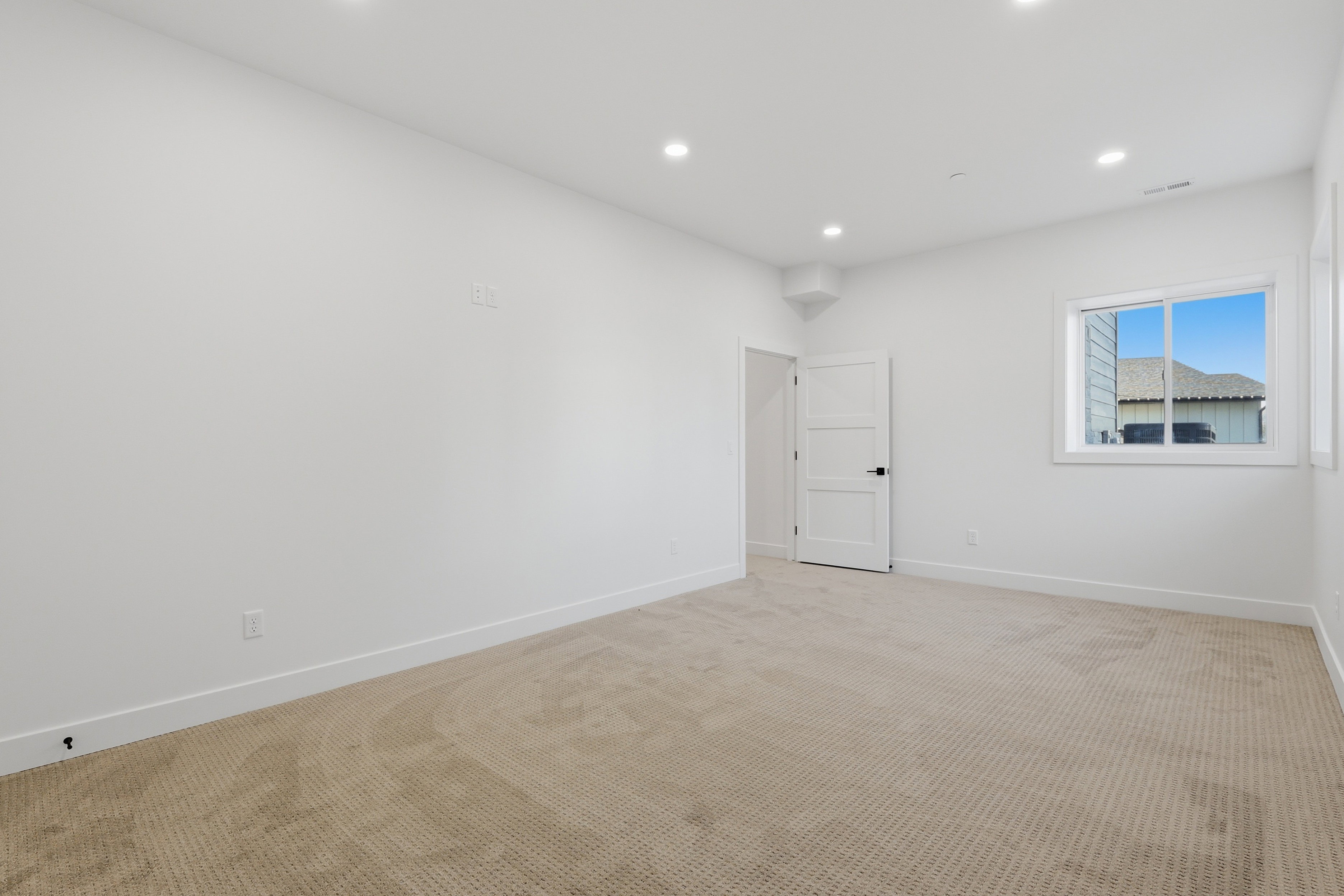Spacious interior room featuring light-toned carpeting, white walls, recessed ceiling lighting, a contemporary white door, and a window providing natural light