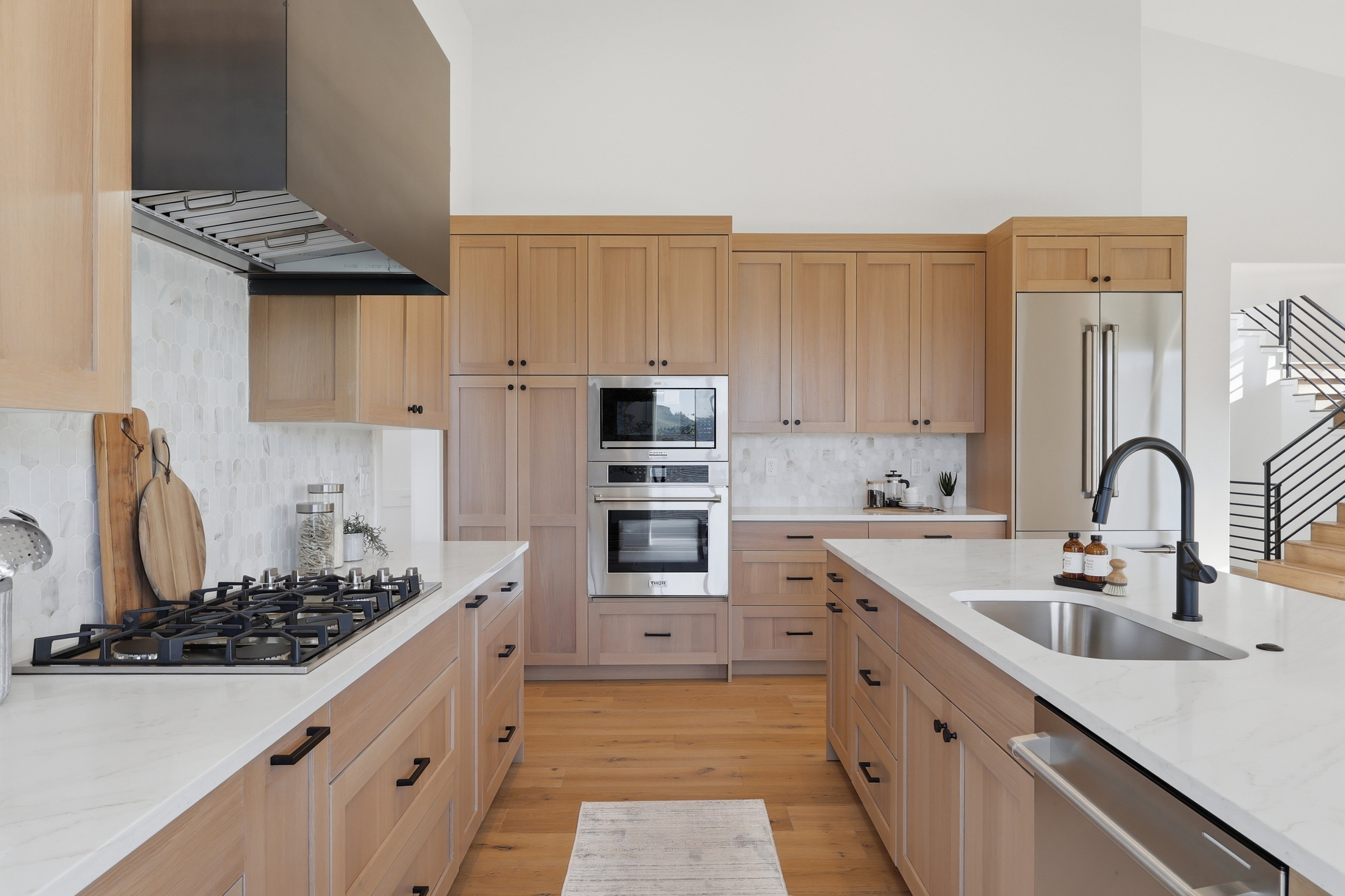 Gourmet kitchen featuring light wood-finish cabinetry with matte black hardware, a large central island with an undermount sink and gooseneck faucet, integrated stainless steel appliances, and a marble-finish tile backsplash
