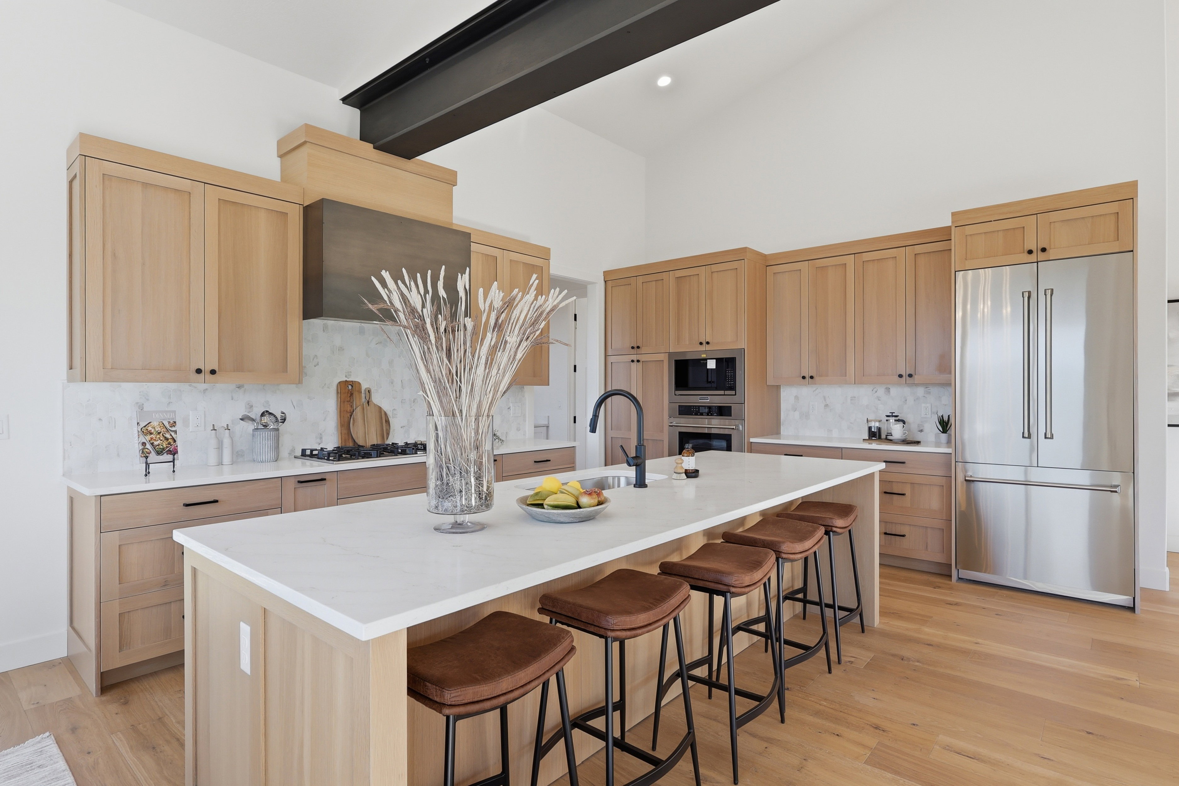 Gourmet kitchen featuring a large central island with seating, stainless steel appliances, light wood cabinetry, white countertops, and a striking exposed ceiling beam