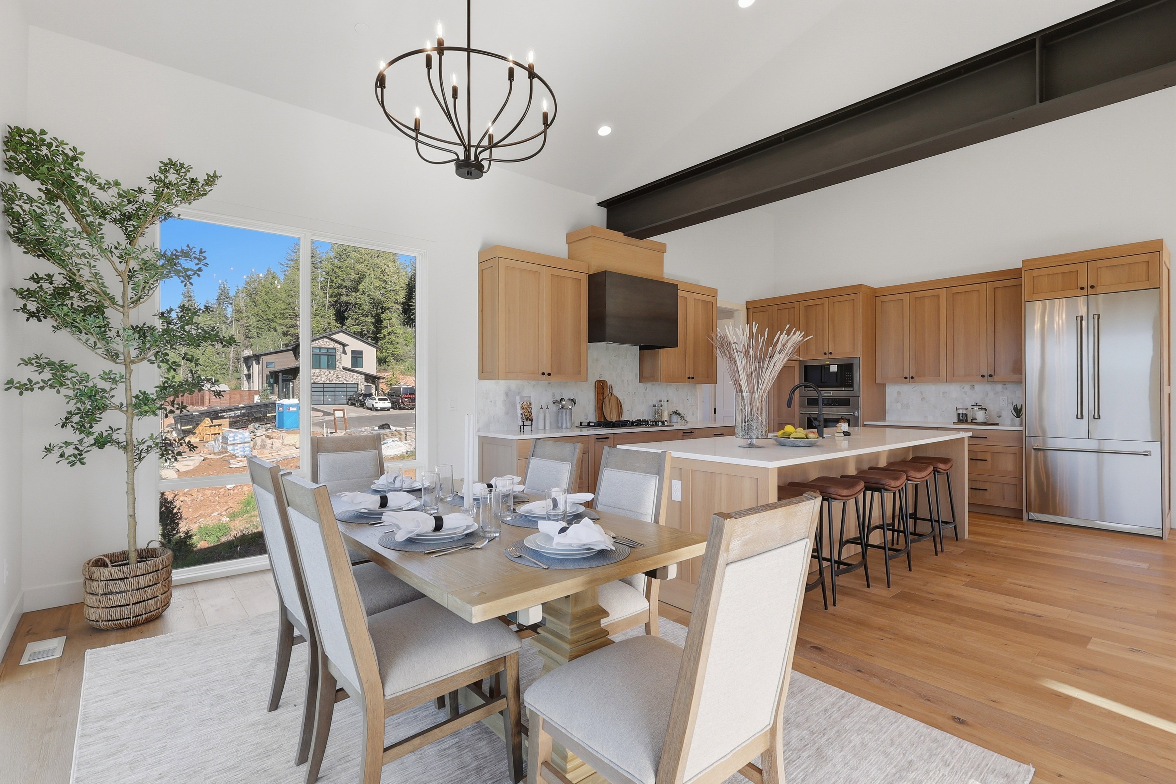 Open-concept kitchen and dining area featuring light wood-finish flooring, a vaulted ceiling with an exposed steel beam, and a contemporary chandelier