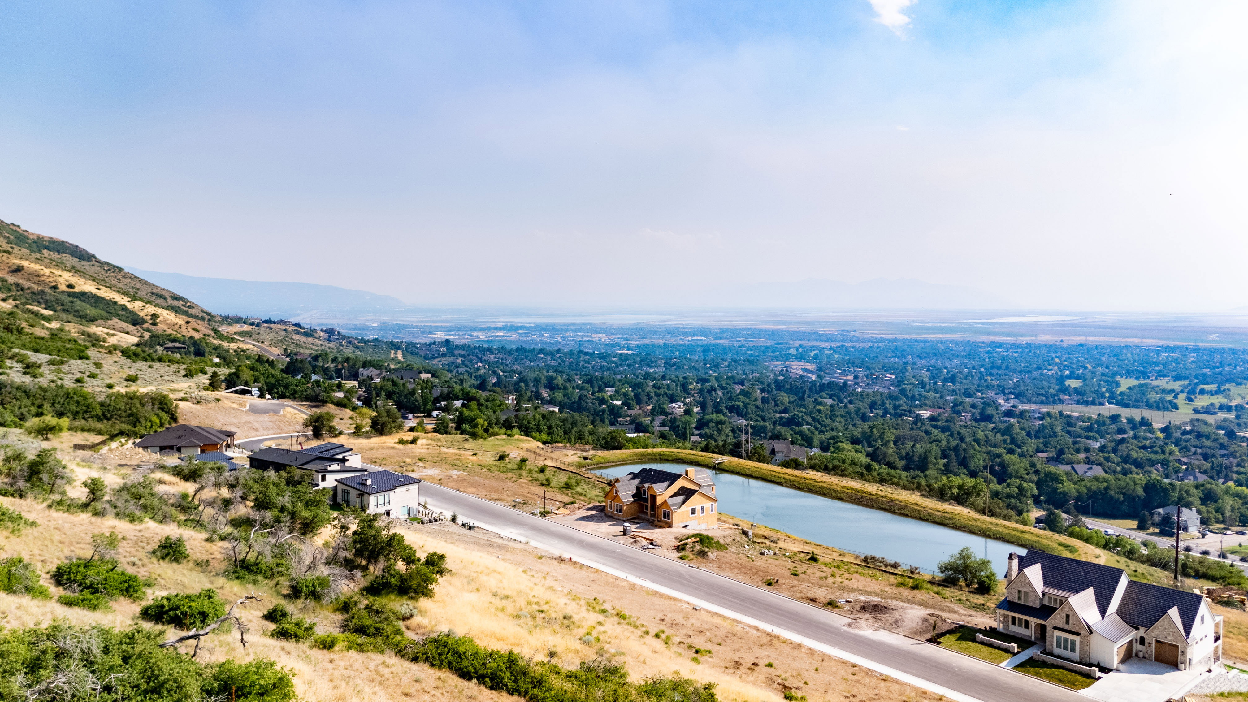 Hillside property featuring a constructed pond, paved roadway, and distant city landscape