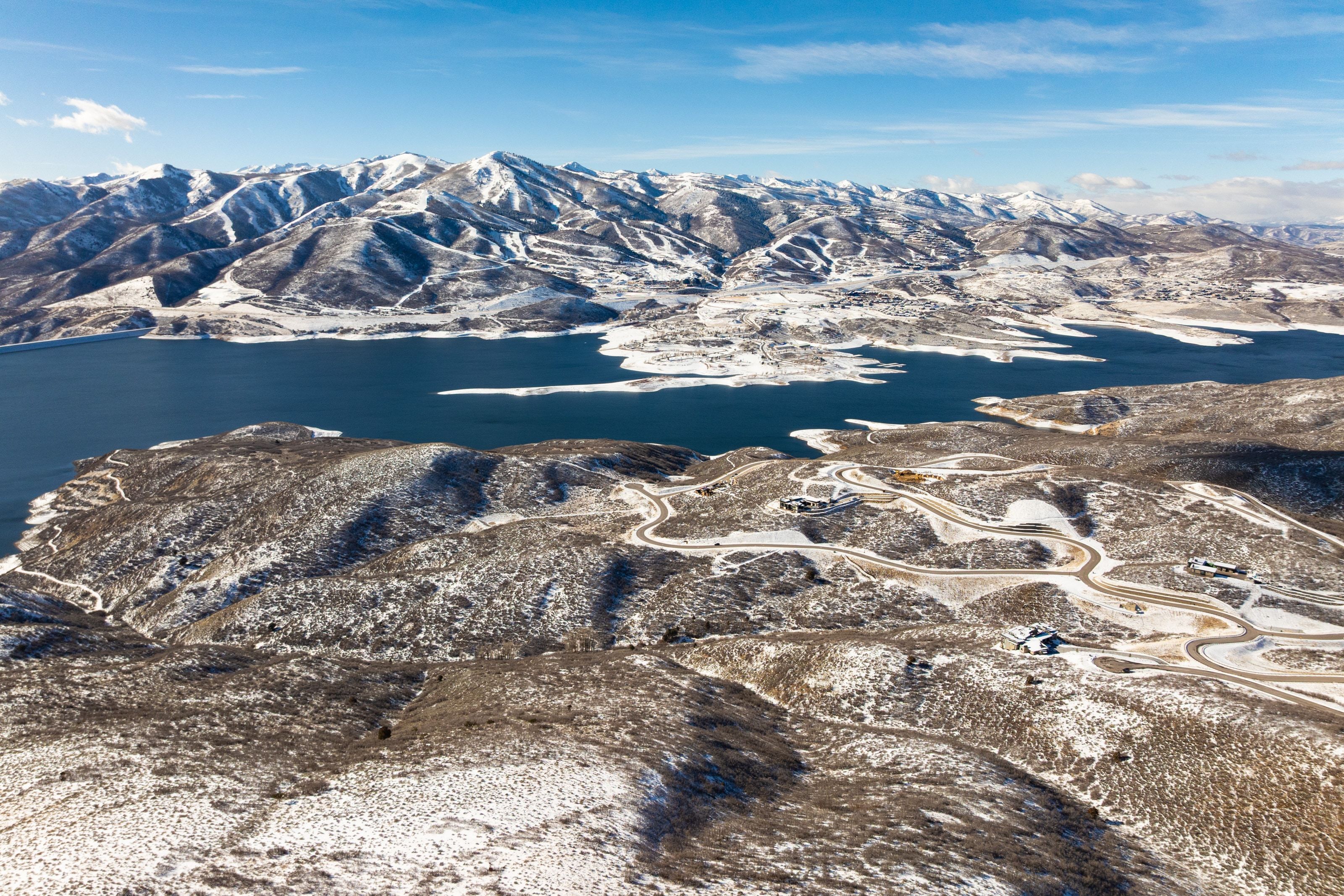 Expansive mountain landscape featuring a large lake, snow-dusted terrain, and winding roads