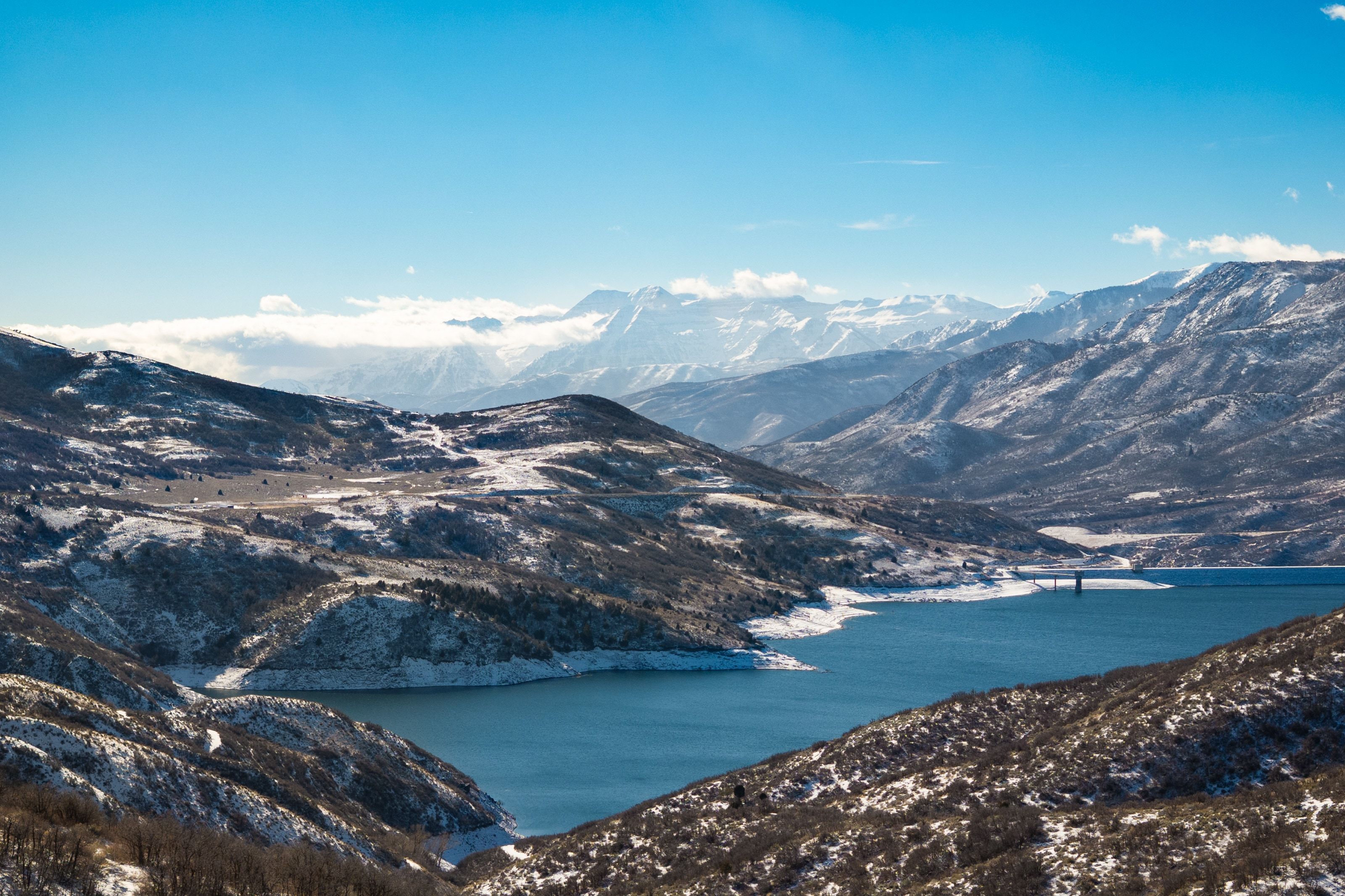 Expansive mountain landscape with a prominent body of water, snow-dusted hillsides, and distant snow-capped peaks under a clear blue sky