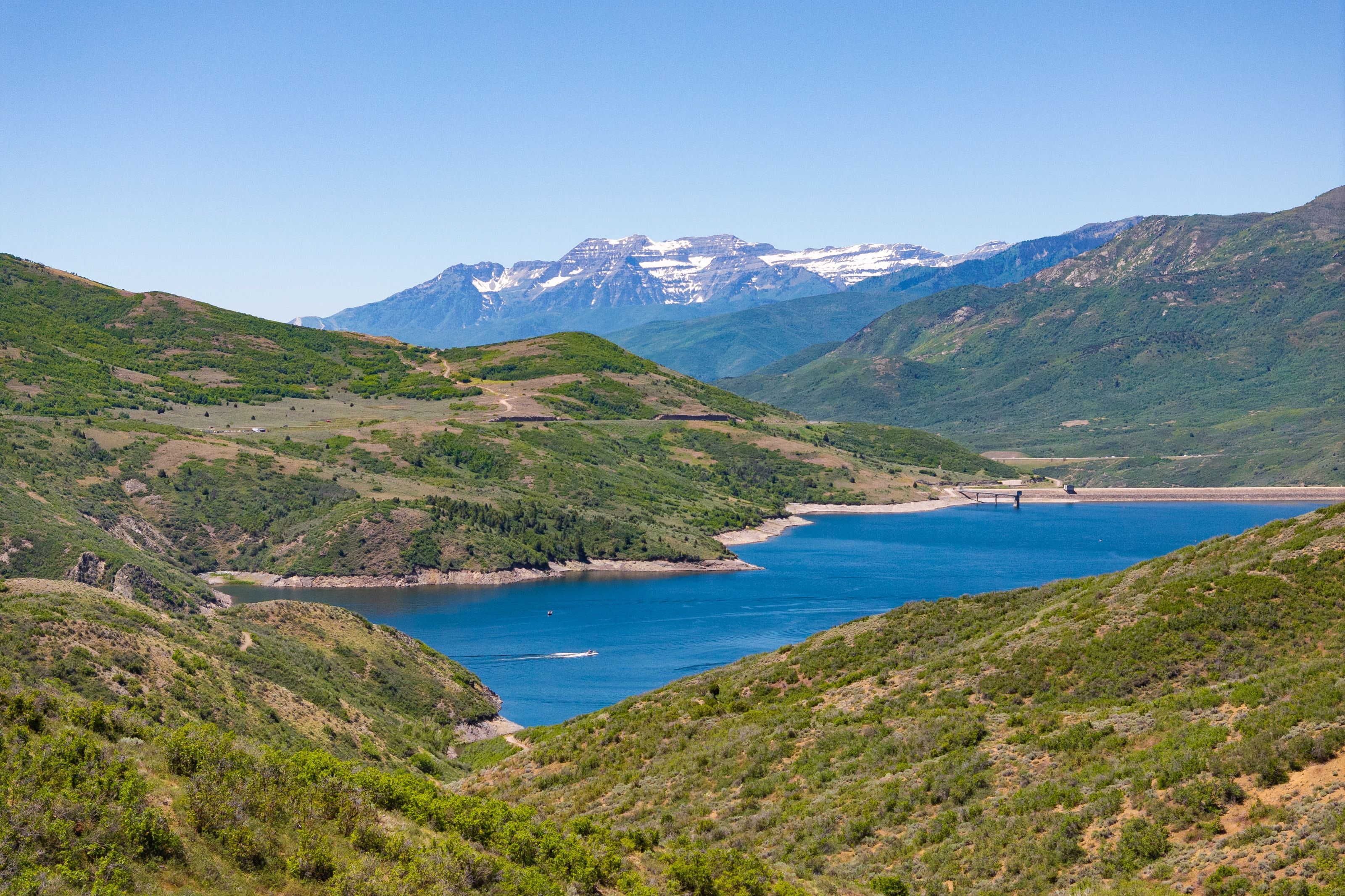 Expansive lake and mountain landscape featuring a deep blue lake, lush green hillsides, and snow-capped peaks in the distance