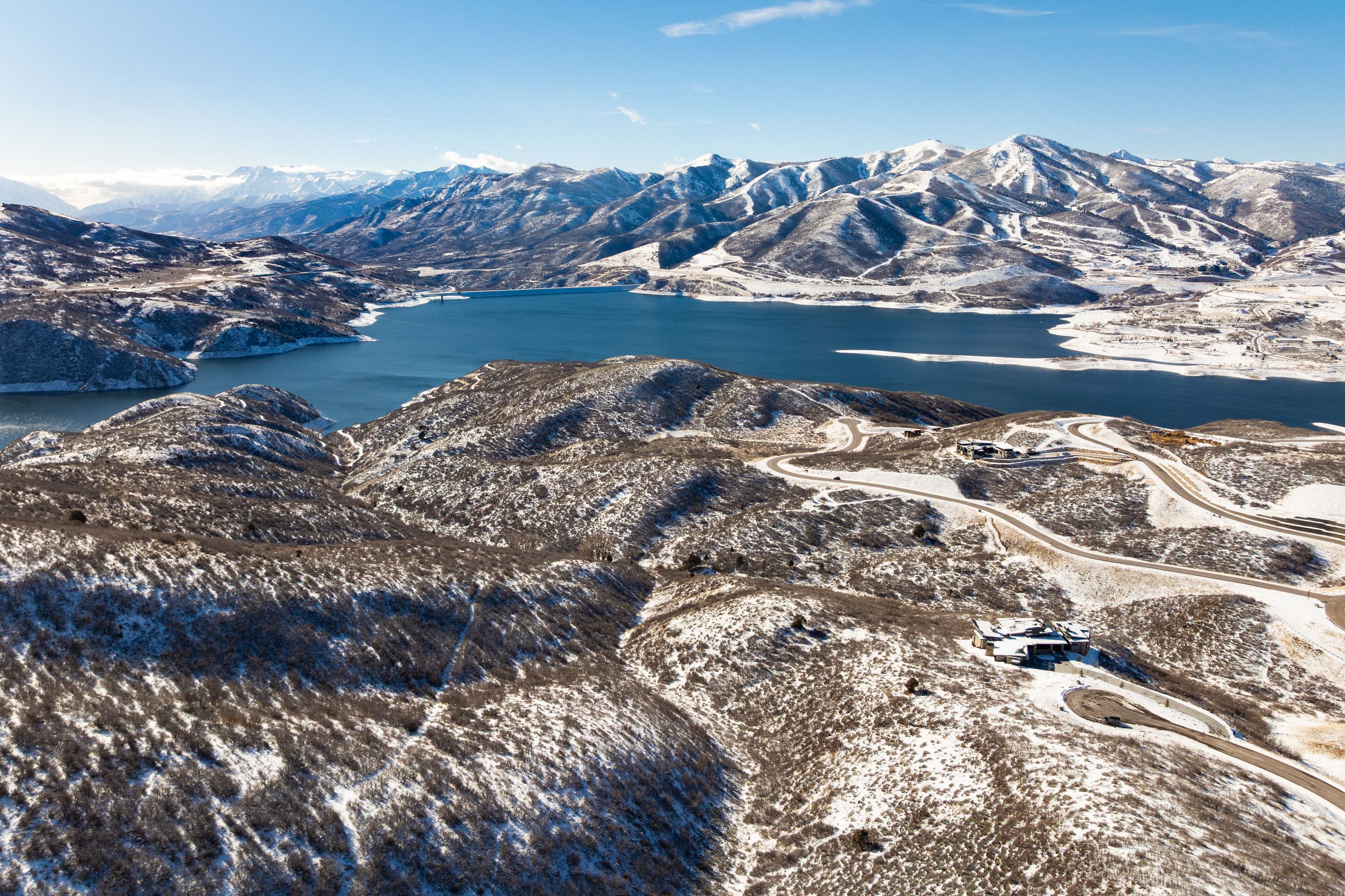 Expansive mountain landscape featuring a large body of water, snow-dusted terrain, and winding roads