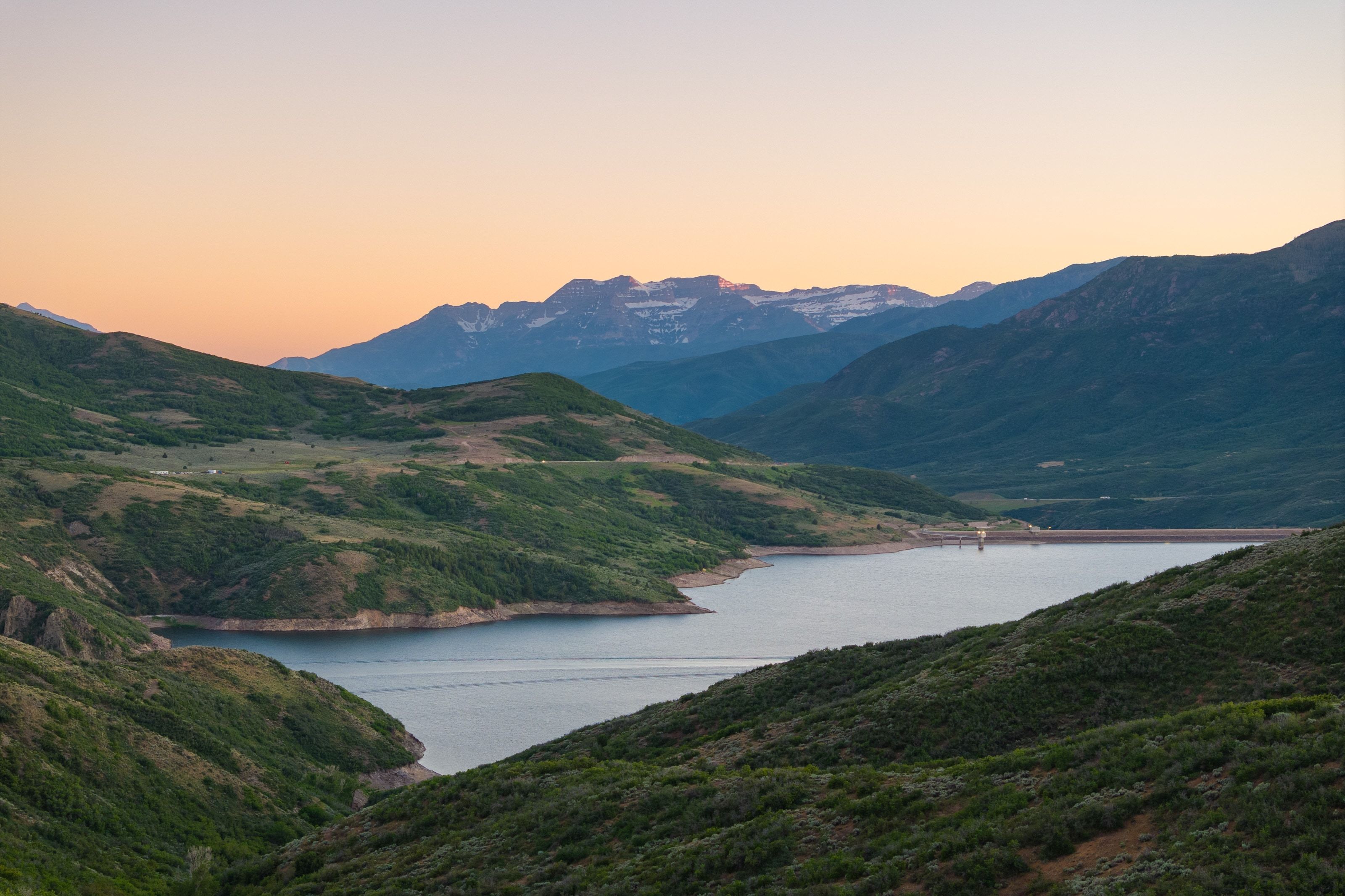 Expansive mountain landscape featuring a large body of water, rolling hillsides, distant snow-capped peaks, and a clear horizon