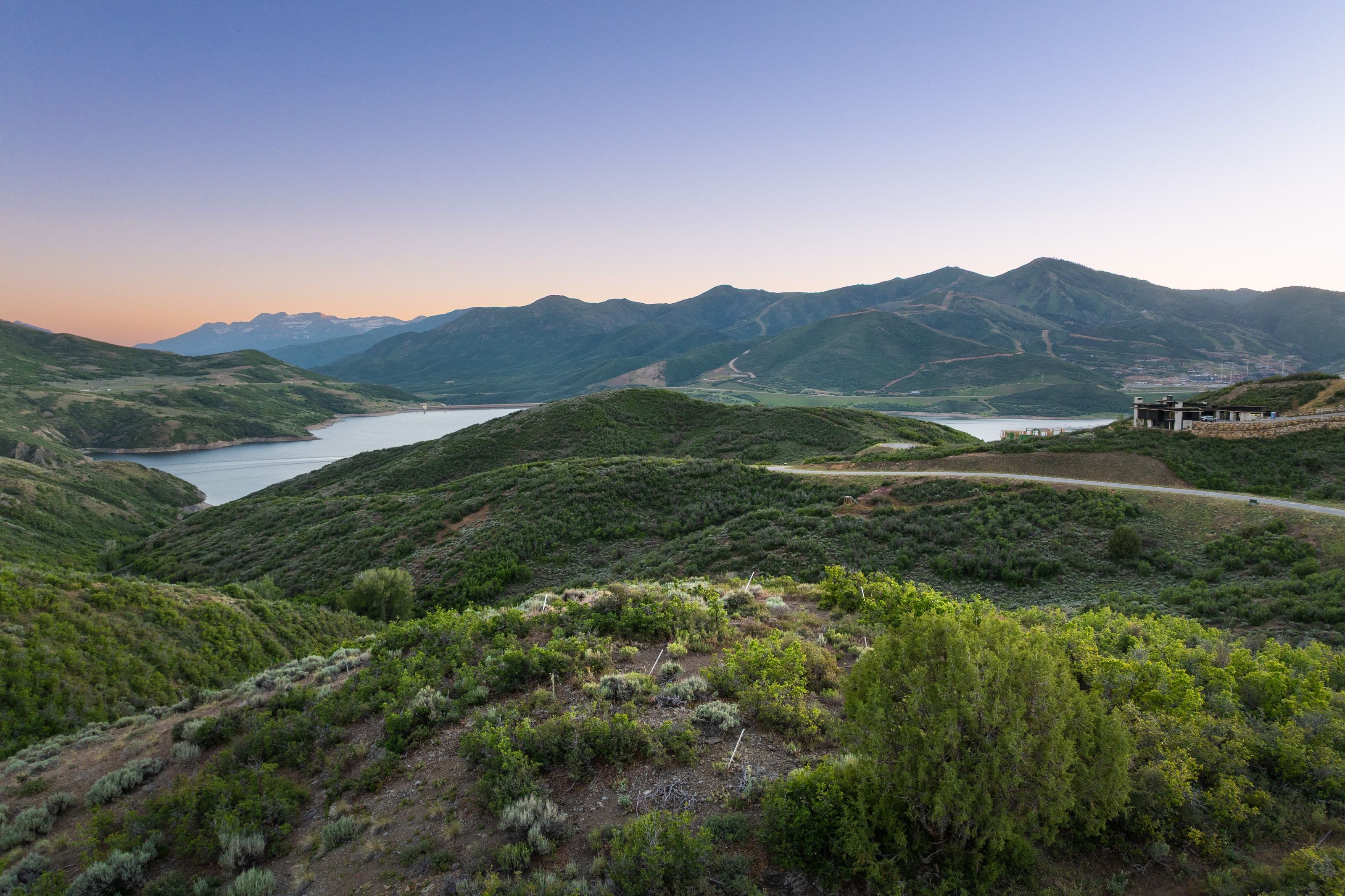 Expansive mountain terrain with a prominent body of water, distant mountain peaks, a winding road, and a contemporary structure with a flat roofline