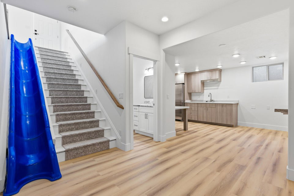 Carpeted staircase with a clear blue slide, wood-finish flooring, and a kitchen featuring light wood cabinetry, a stainless steel refrigerator, and a stainless steel sink