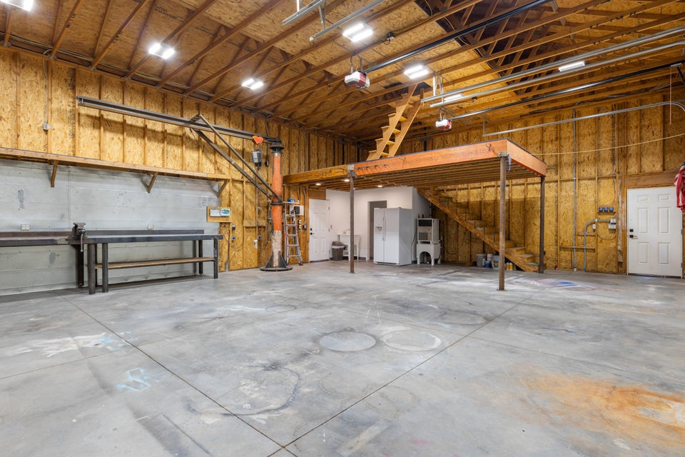 Expansive concrete flooring in a large utility space featuring a built-in loft platform with wood stairs, exposed wood-beam ceiling, and multiple wall-mounted light fixtures