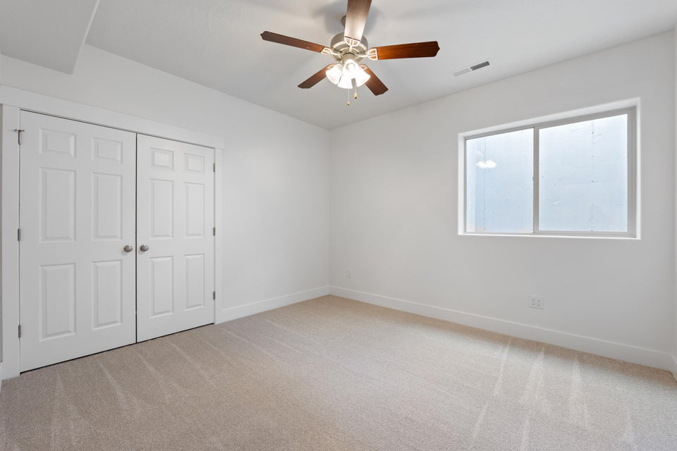 Neutral-toned carpeted room featuring a ceiling fan with integrated lighting, a double-door closet with paneling, a large window with white trim, and white baseboards