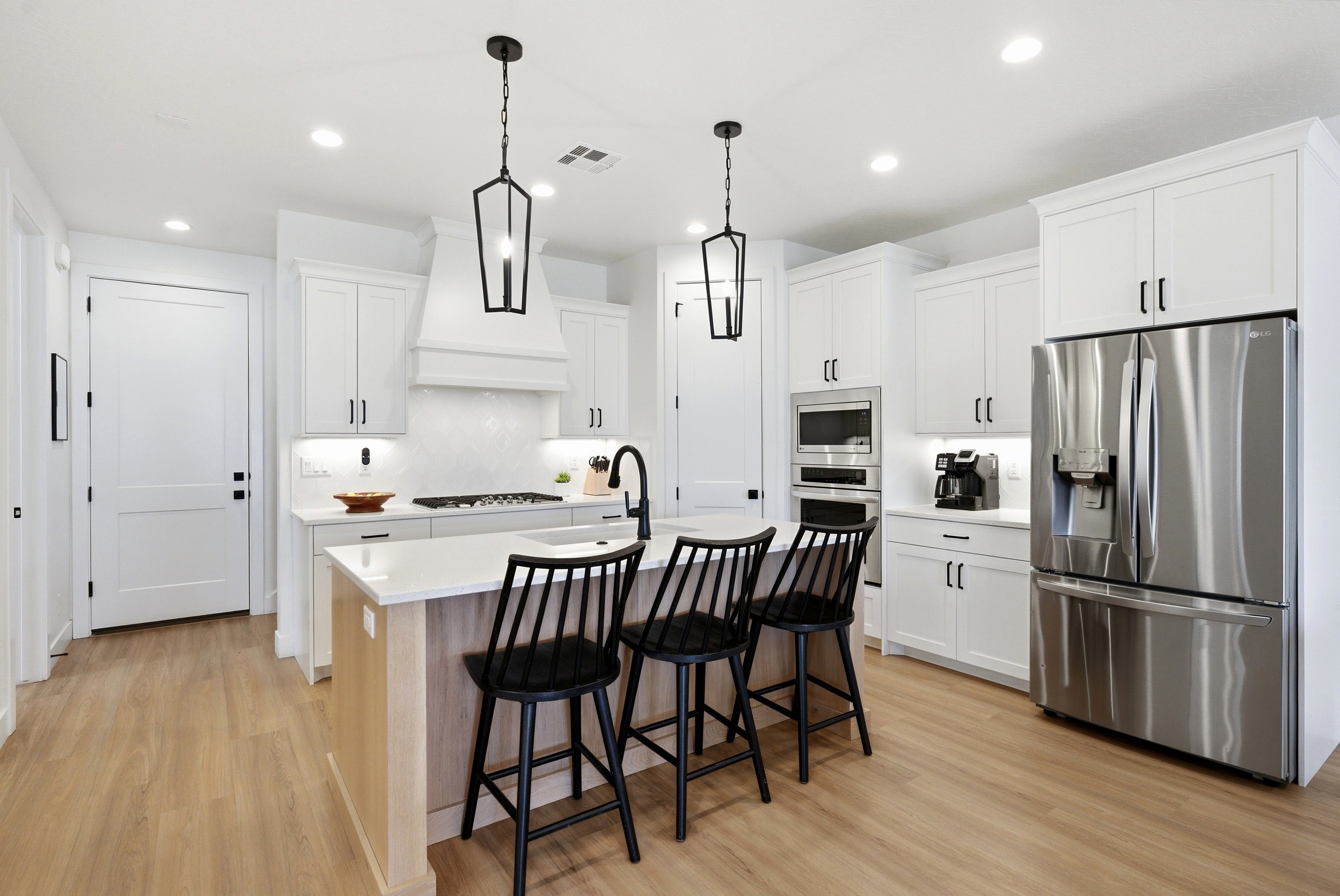 Contemporary kitchen featuring a large center island with seating, stainless steel appliances, white cabinetry with black hardware, wood-finish flooring, and recessed and pendant lighting