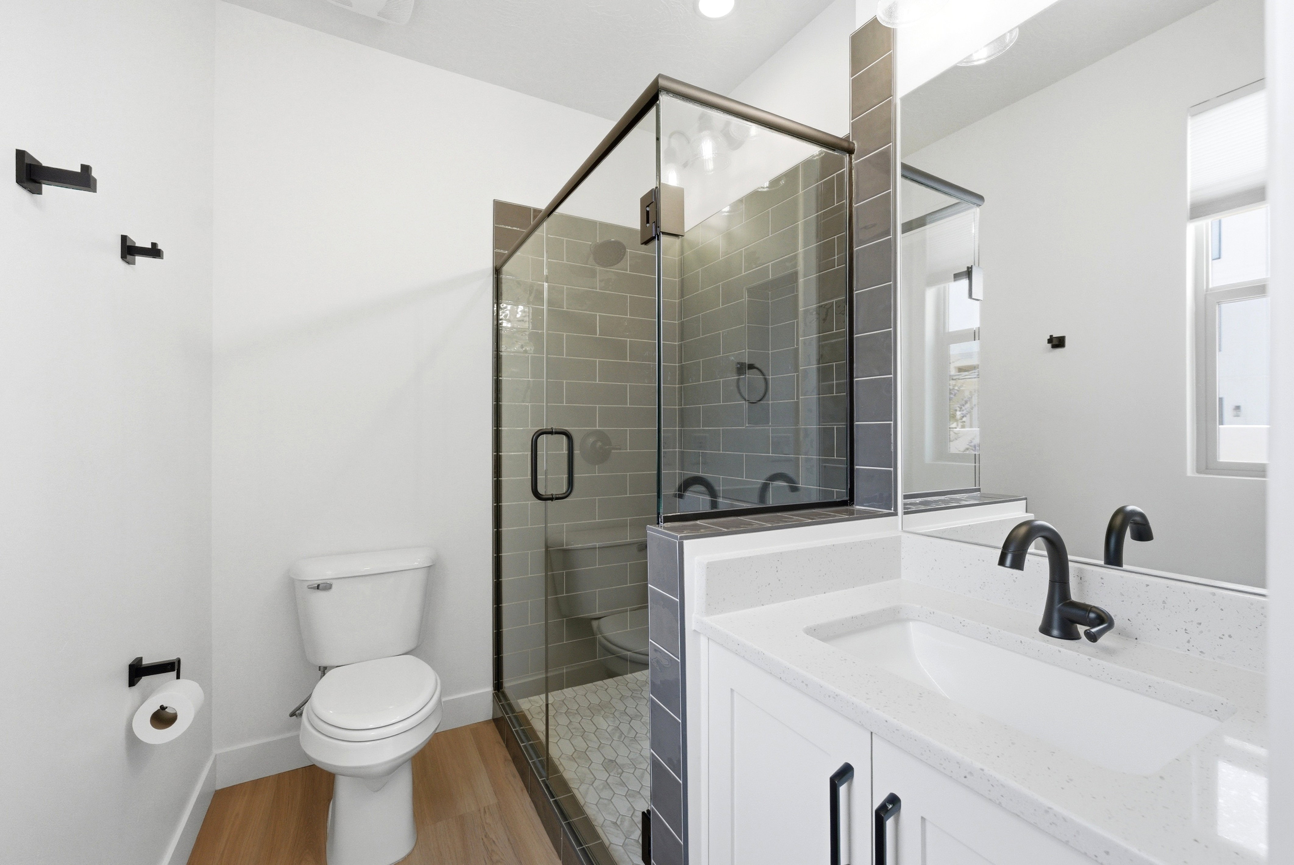 Bathroom featuring a glass-enclosed shower with subway tile surround, white vanity with integrated sink and stone-finish countertop, wood-finish flooring, and matte black fixtures
