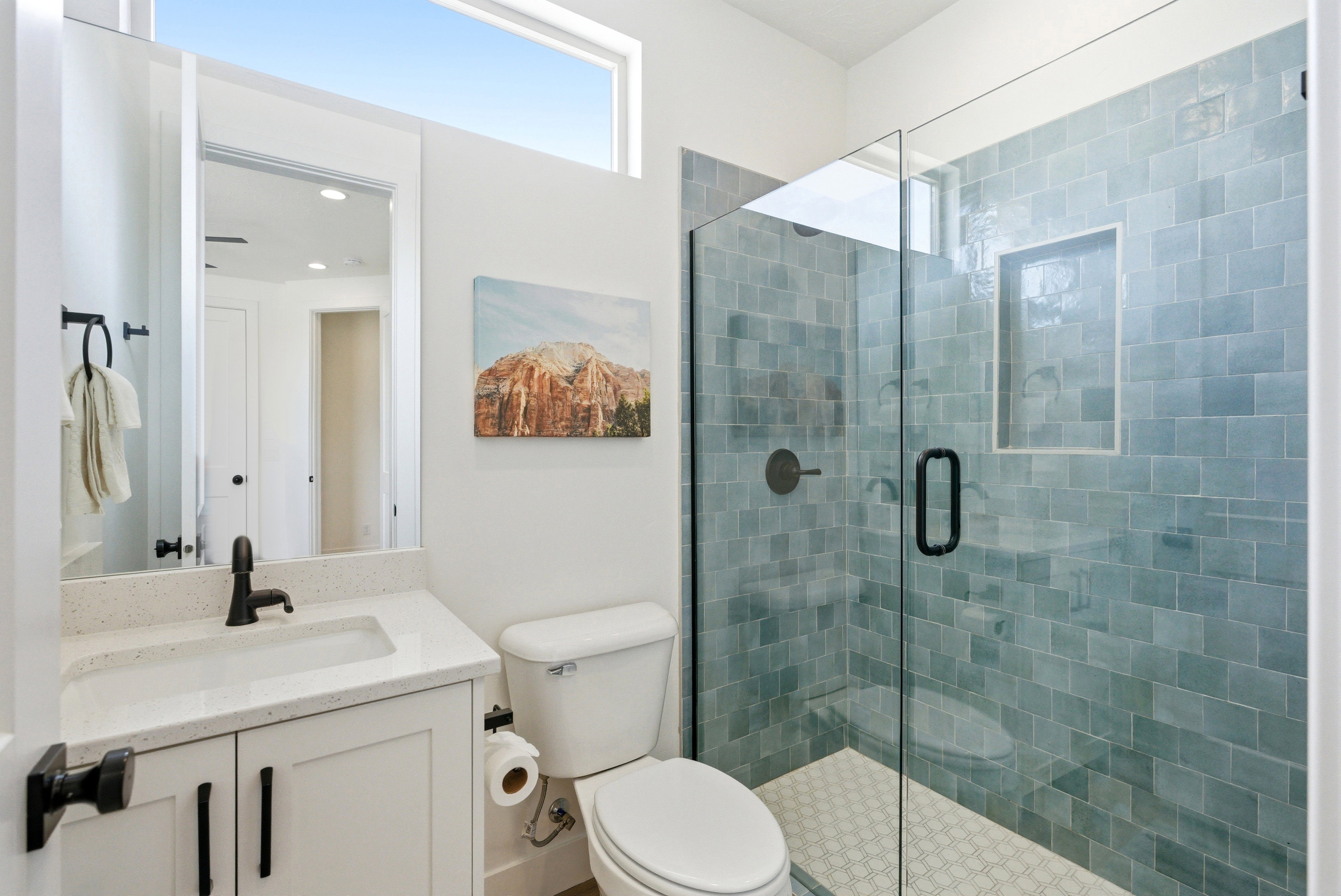 Contemporary bathroom featuring a clear glass shower enclosure with blue-grey subway tile, a white vanity with a light speckled countertop, and matte black fixtures