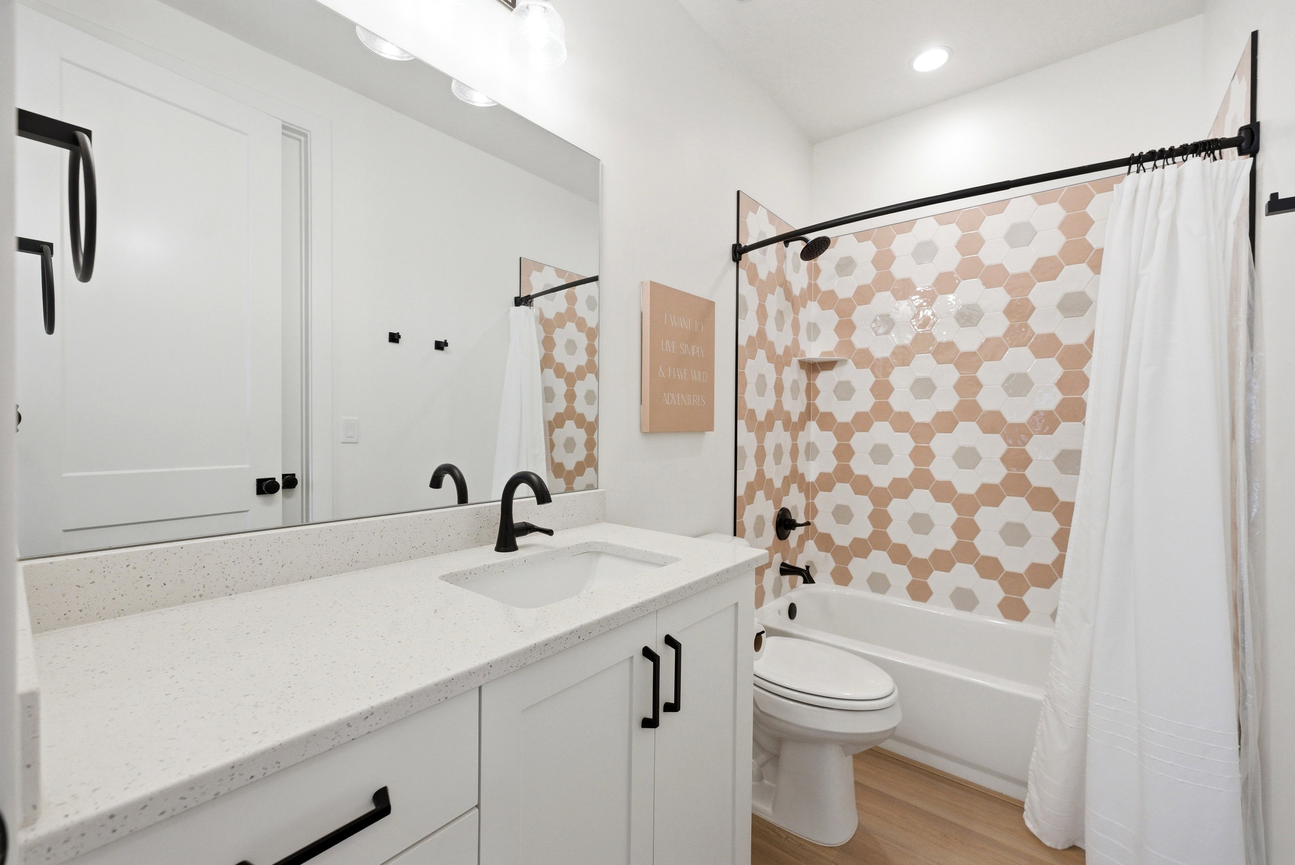 Bathroom featuring a white vanity with a light-toned countertop, matte black fixtures, wood-finish flooring, a full-width mirror, and a tub/shower combination with decorative hexagonal tiling