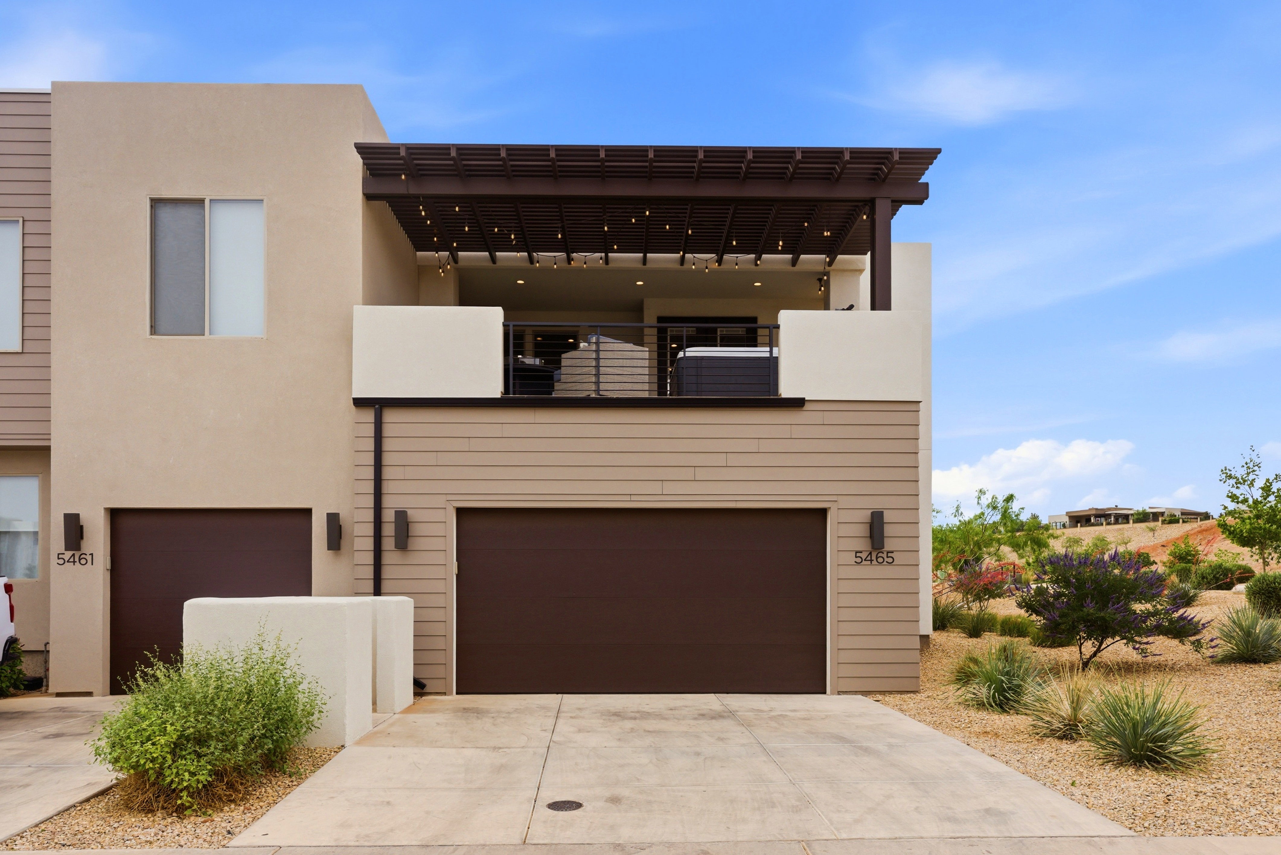 Multi-level exterior featuring a ground-floor garage with a brown door, an upper-level balcony with a wood pergola, and desert landscaping