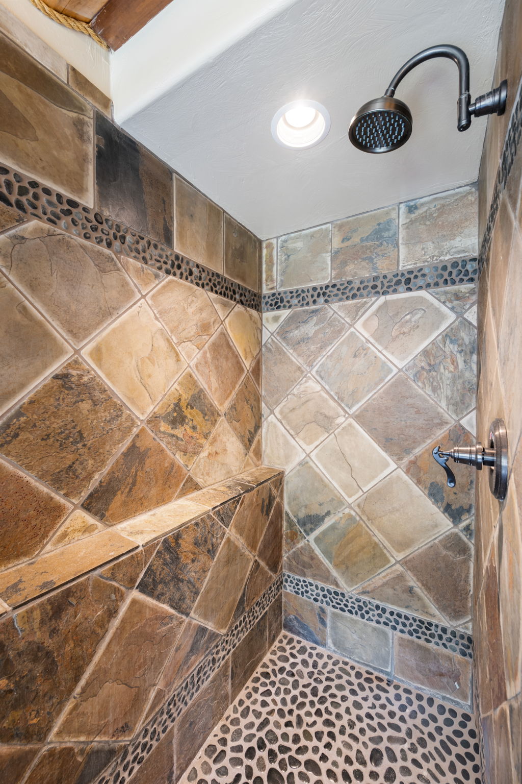 Spacious shower stall featuring natural stone tile walls in a diagonal pattern, pebble tile flooring, a built-in shower bench, and a bronze finish rain shower head